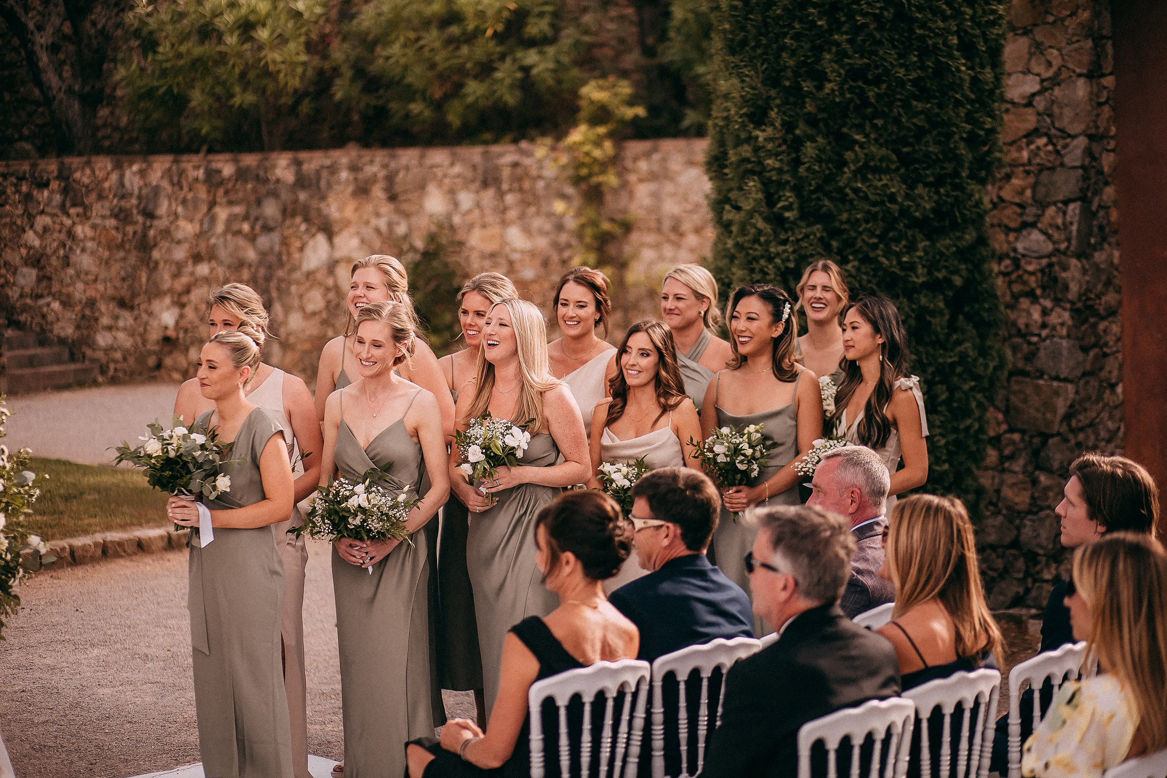 A group of bridesmaids standing together, all wearing elegant sage-green dresses, smiling and holding bouquets during an outdoor ceremony.