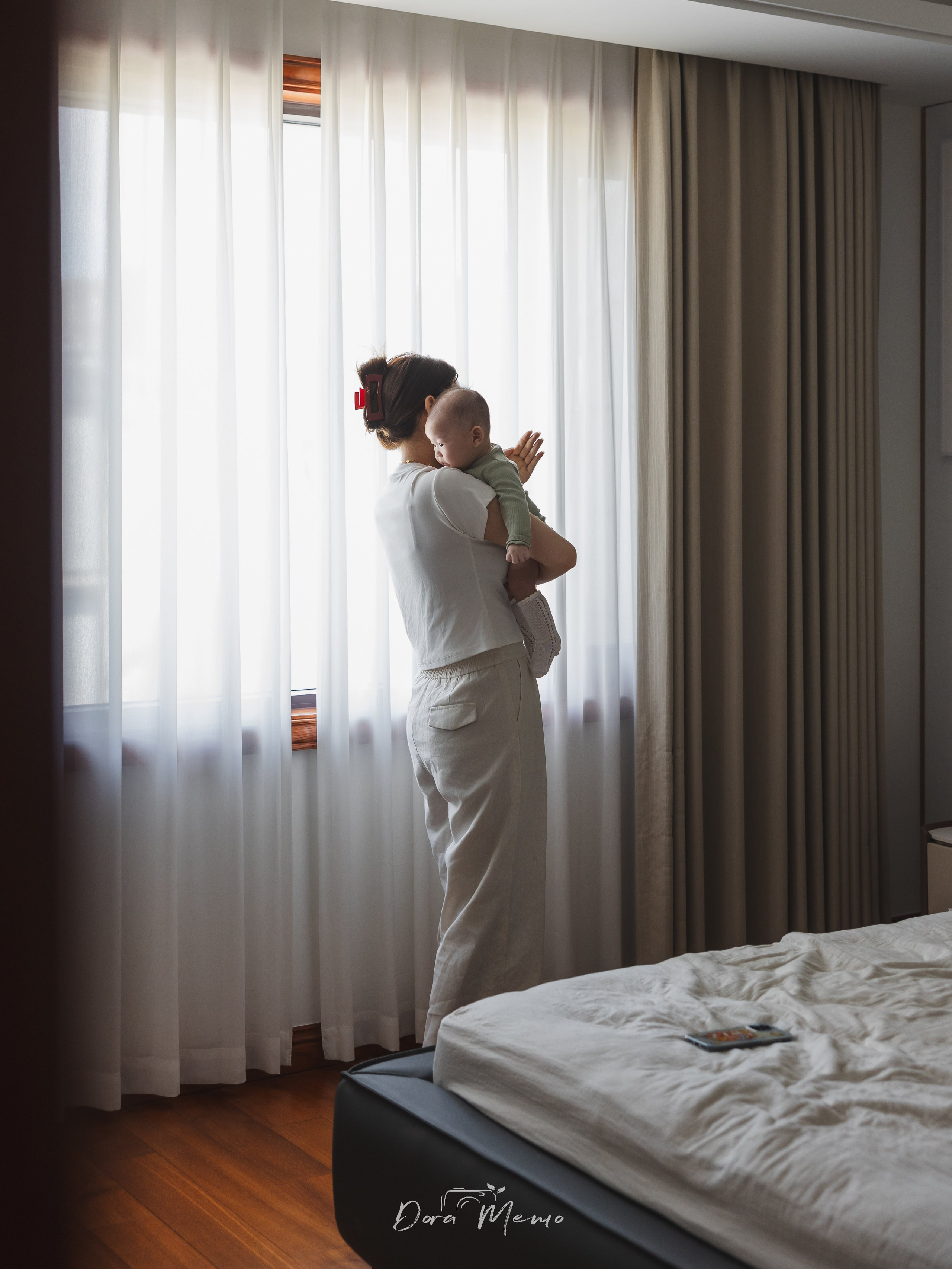  A mother holds her newborn by the window, both gazing at the soft light outside