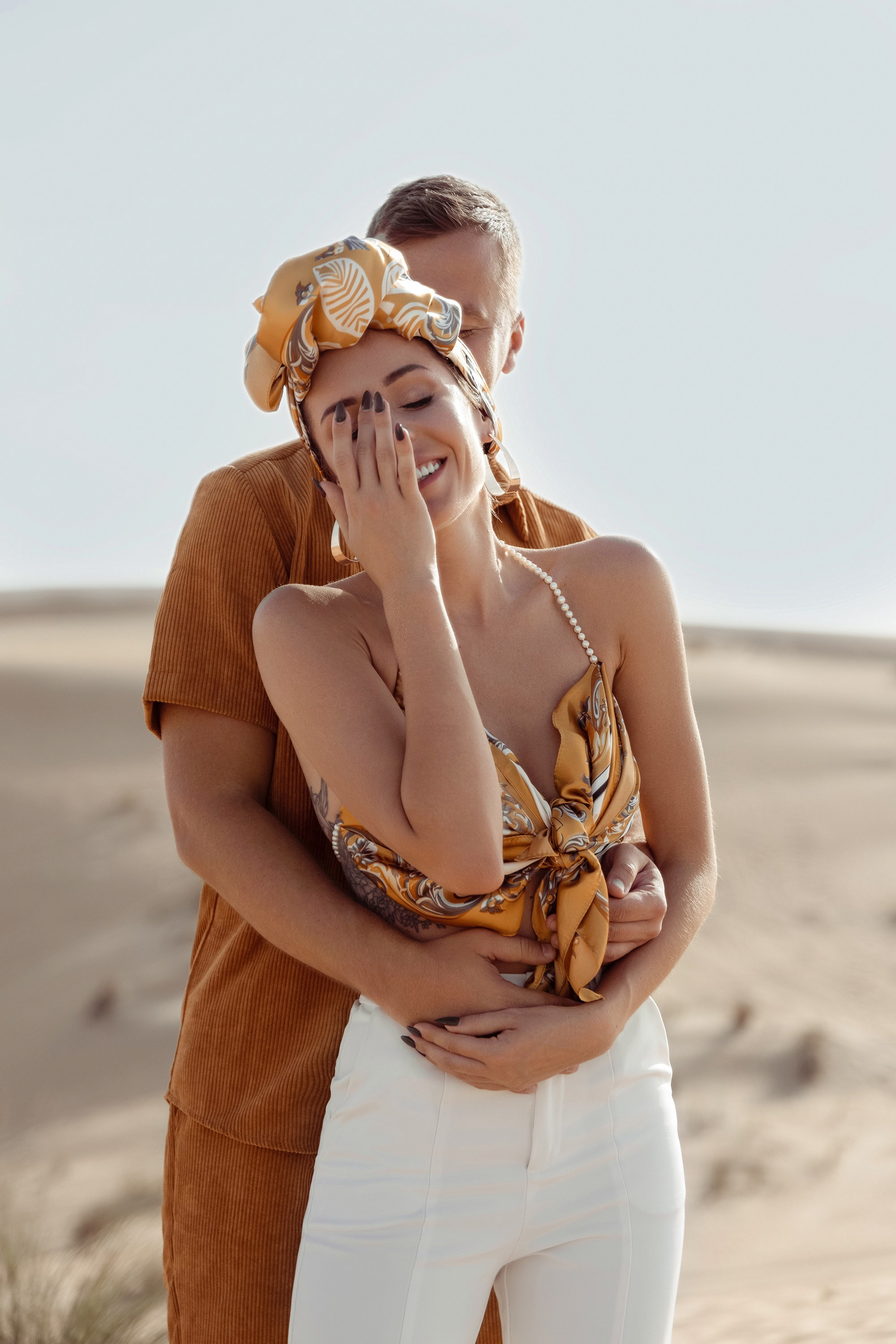 A candid shot of a couple in the desert, sharing a tender hug and playful laughter as the sun casts long shadows on the sand. Dubai, United Arab Emirates