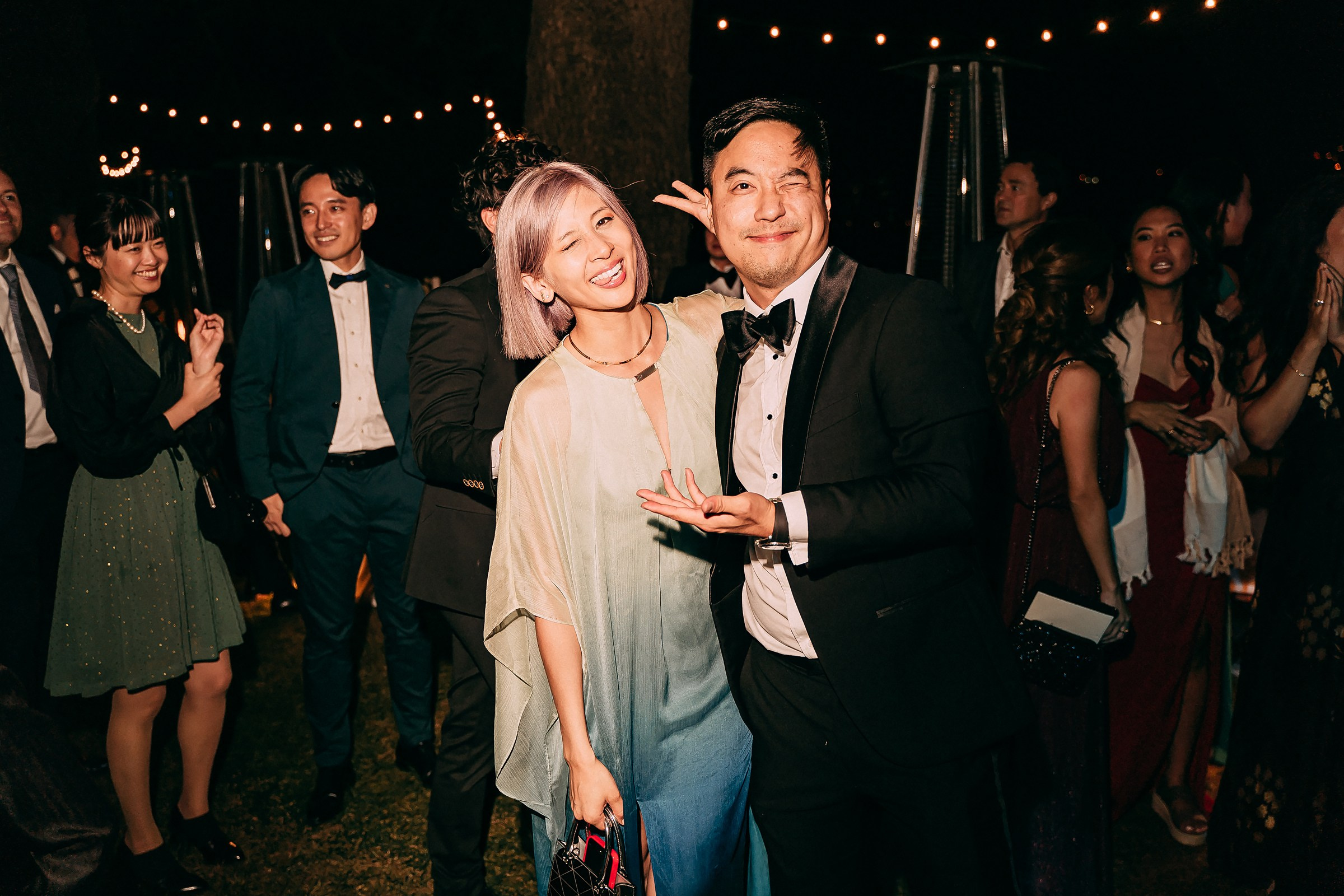 Smiling couple in formal attire poses together outdoors, surrounded by friends in evening wear.