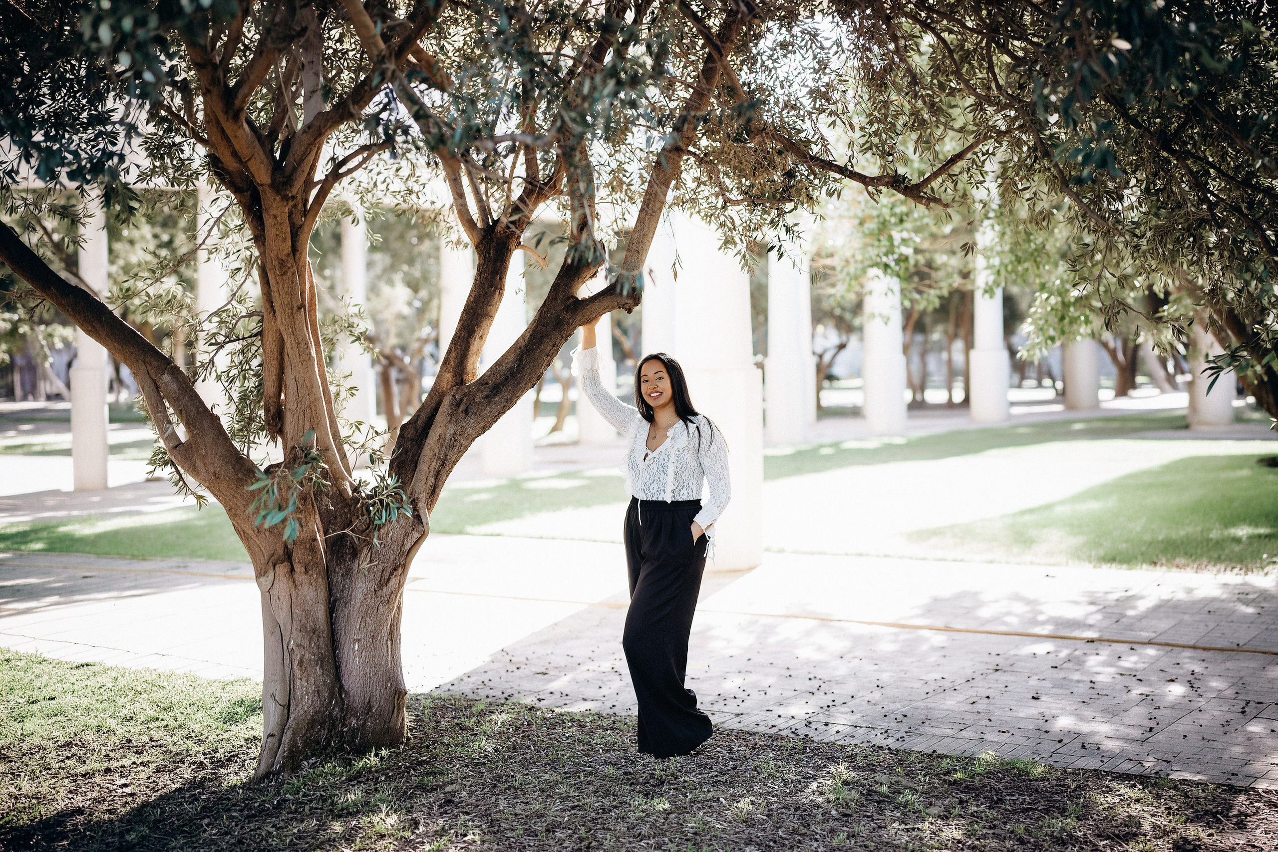 Женская портретная фотосессия в Валенсии, Испания: улыбающаяся женщина стоит у оливкового дерева в солнечном парке.