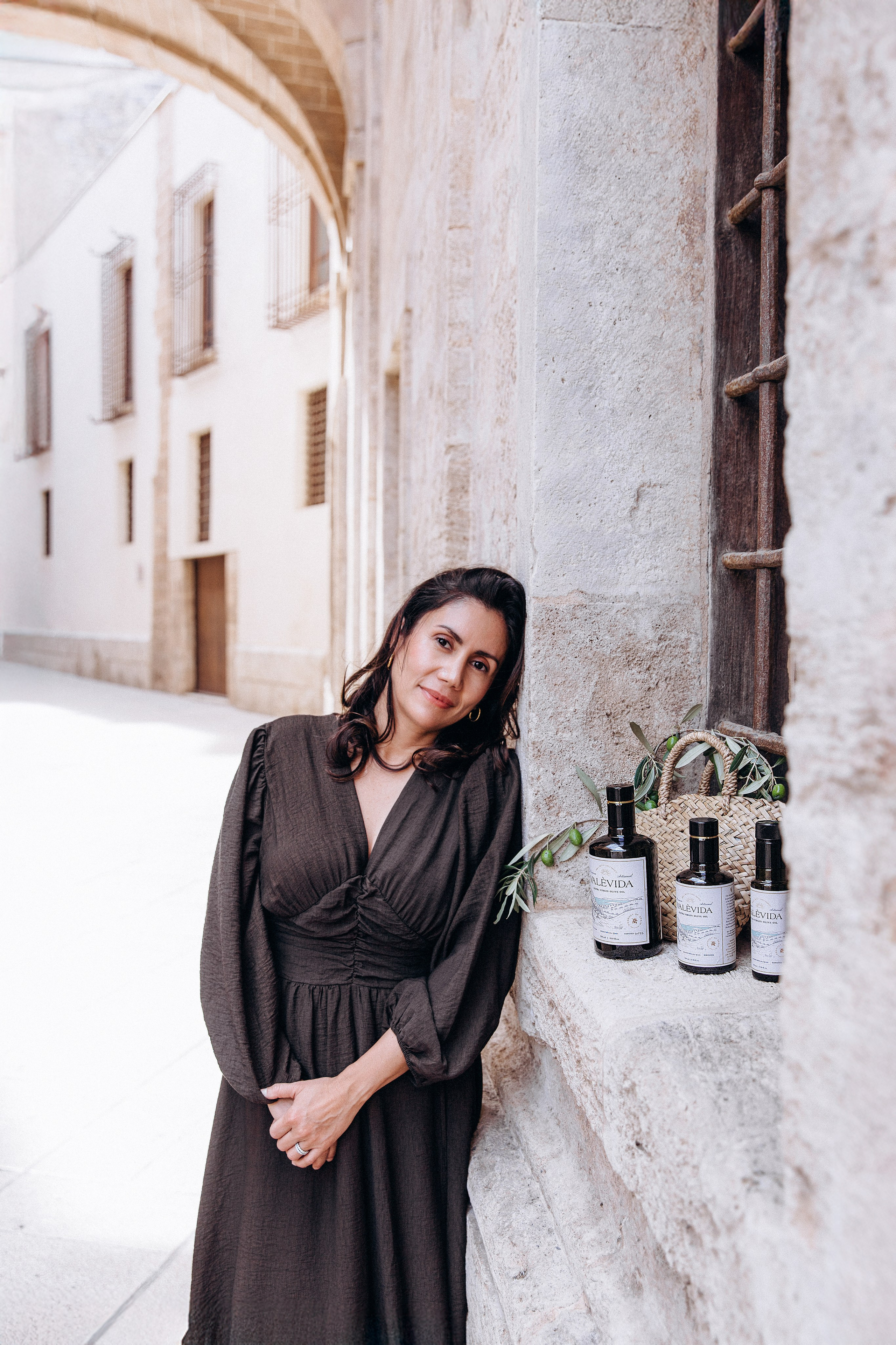 Stylish brand photography in Valencia, Spain, featuring a woman in an elegant black dress standing beside a rustic window ledge displaying artisan product bottles — ideal for showcasing natural and lifestyle brands. Great example of content and product photoshoots in Valencia for businesses seeking authentic Mediterranean visuals.
