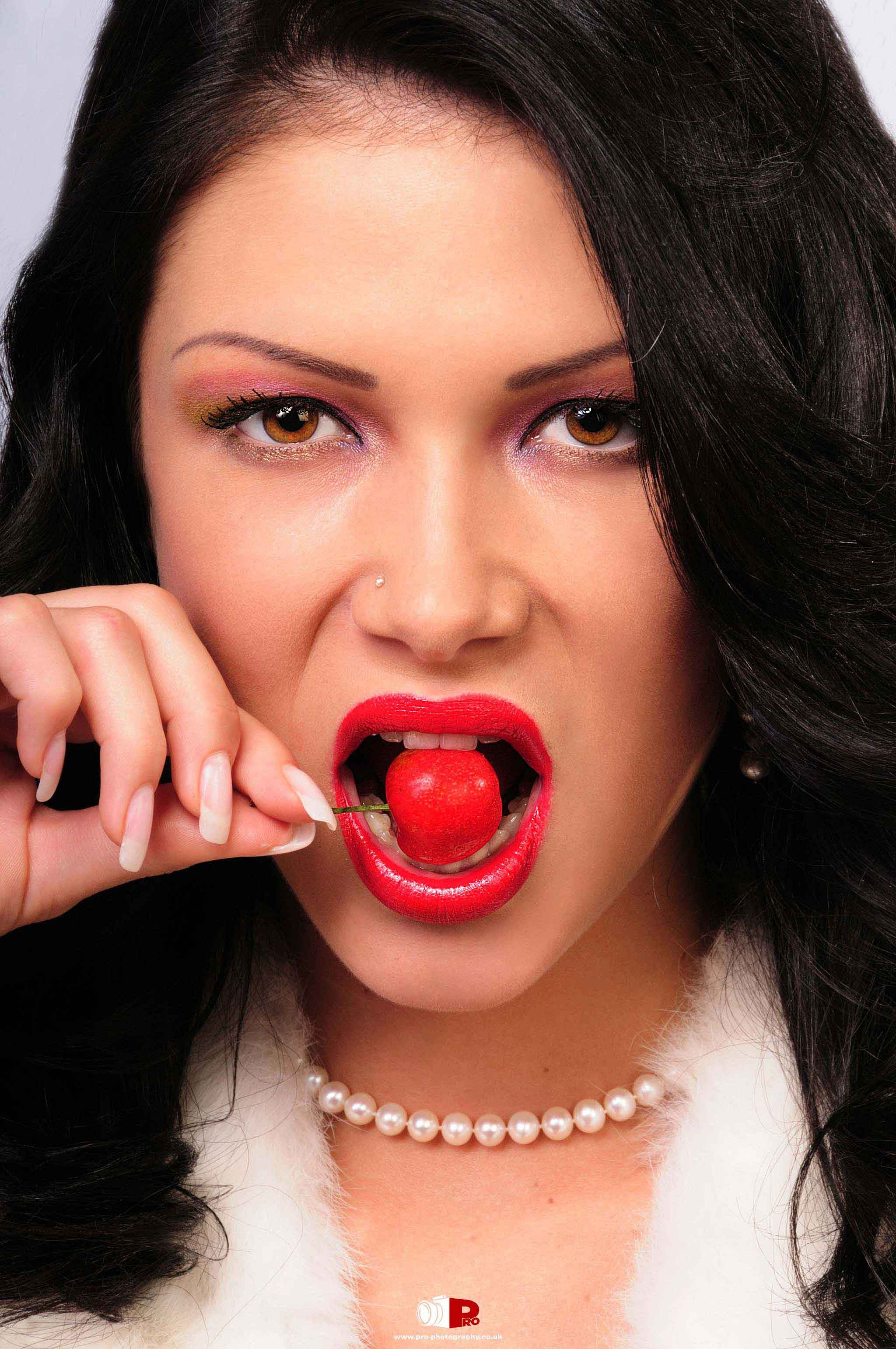 Stylish woman with red lipstick biting a cherry, wearing pearl jewellery and white fur clothing.