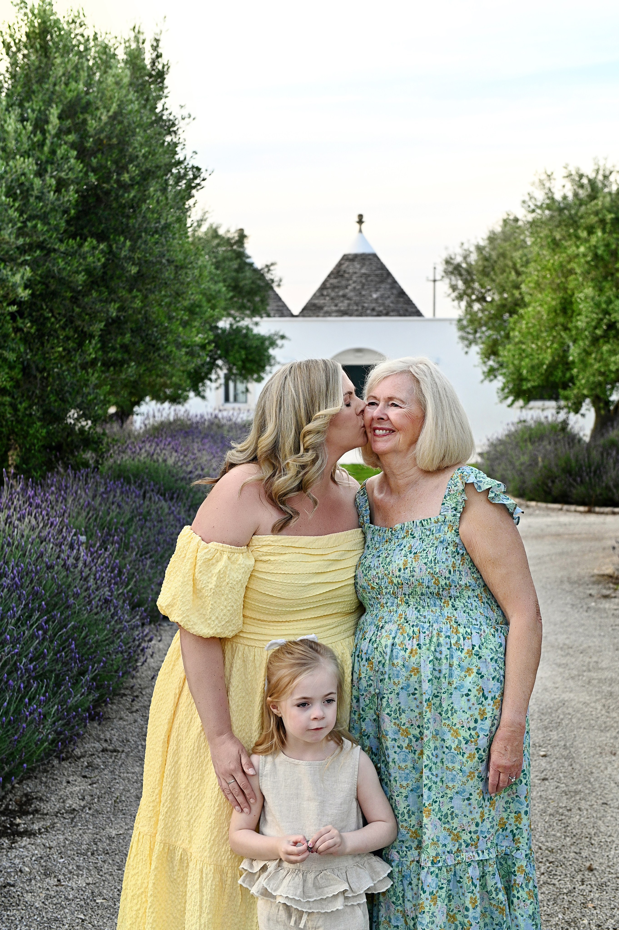 Servizio fotografico di famiglia in Puglia: una donna in abito giallo bacia sulla guancia la sua mamma sorridente, mentre la nipotina è accanto a loro su un sentiero di lavanda con un trullo sullo sfondo.