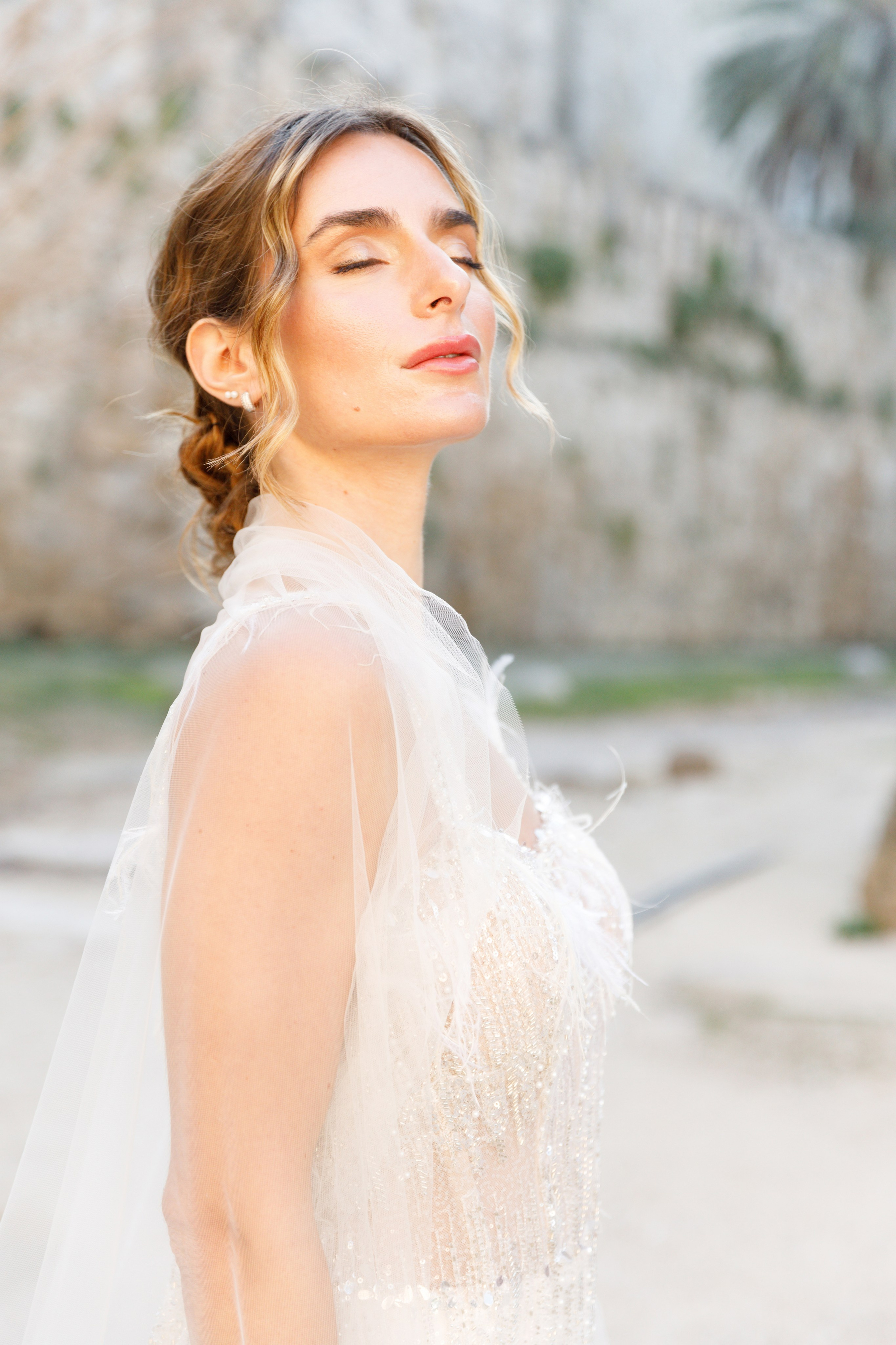 A radiant bride stands in the narrow, sunlit streets of Rhodes' Old Town, her intricate lace wedding dress contrasting beautifully with the weathered stone walls behind her. The editorial-style portrait highlights her serene expression and the timeless elegance of the historic setting, with soft natural light accentuating her features.