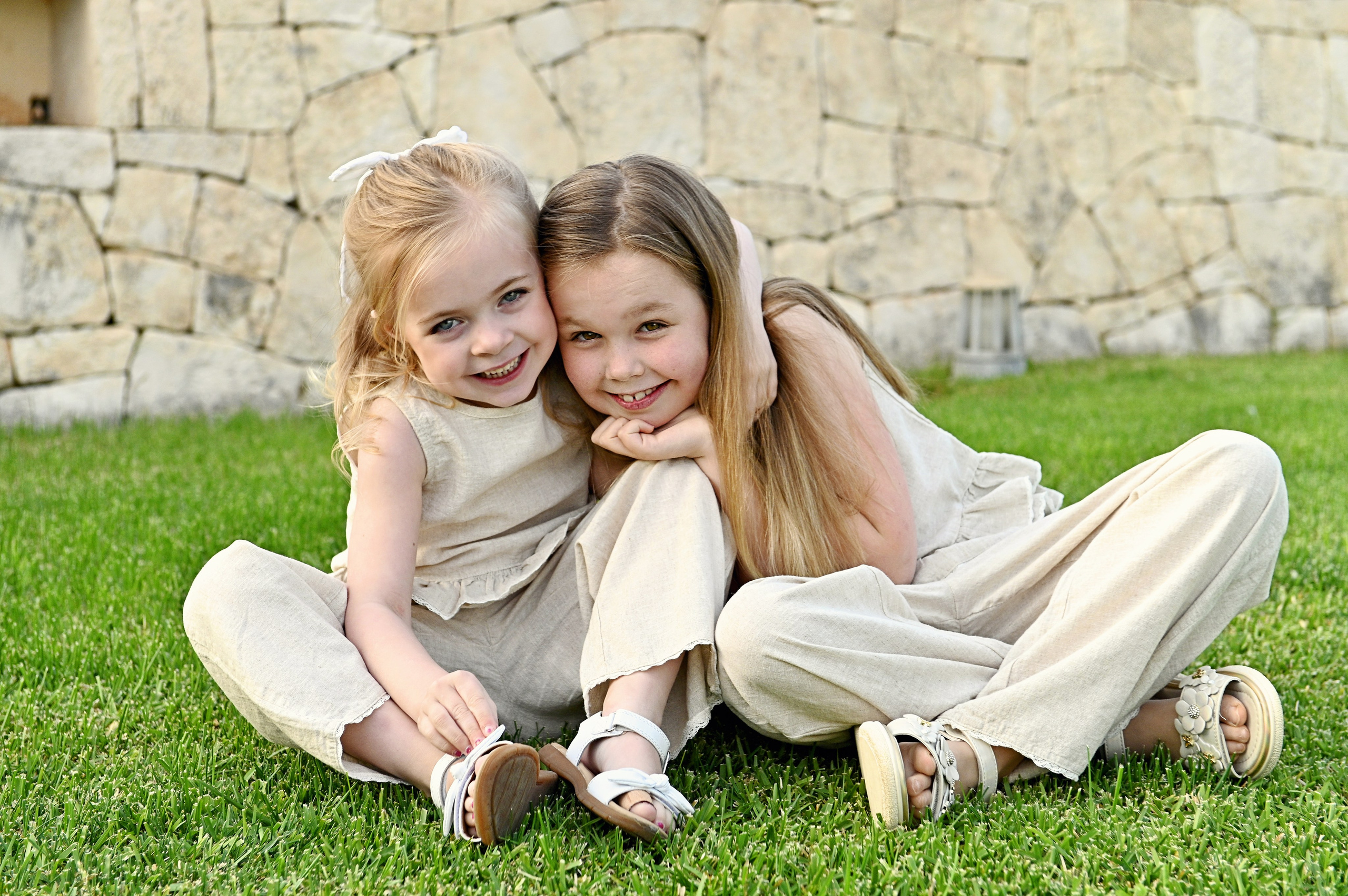 Servizio fotografico per bambini in Puglia: due bambine sorridenti in completi di lino crema sedute e abbracciate sull'erba verde davanti a un muro di pietra.