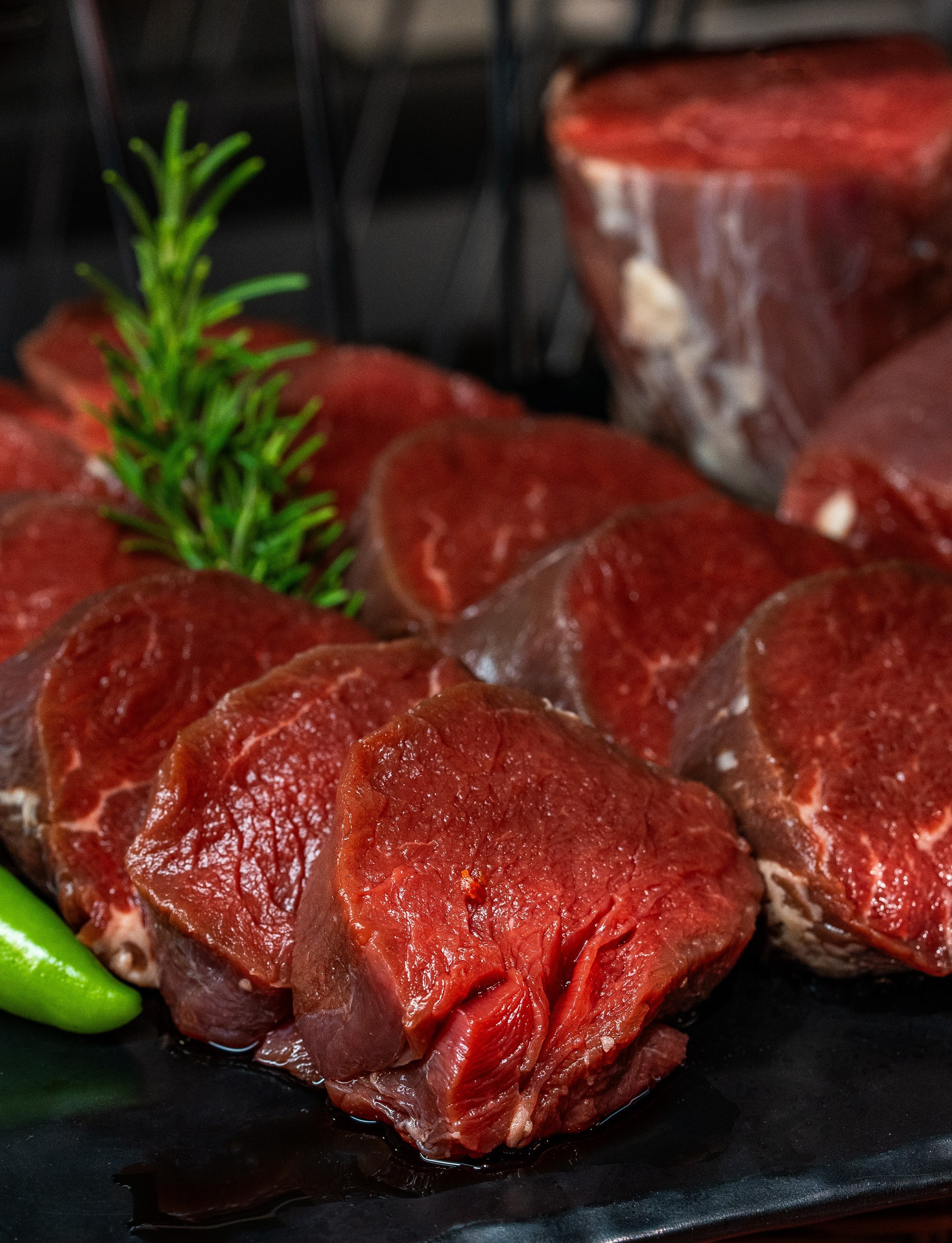 Təzə kəsilmiş, qırmızı, qalın dana filesi (file mignon) dilimləri, bir budaq rozmarin və yaşıl bibər ilə qara fonda.