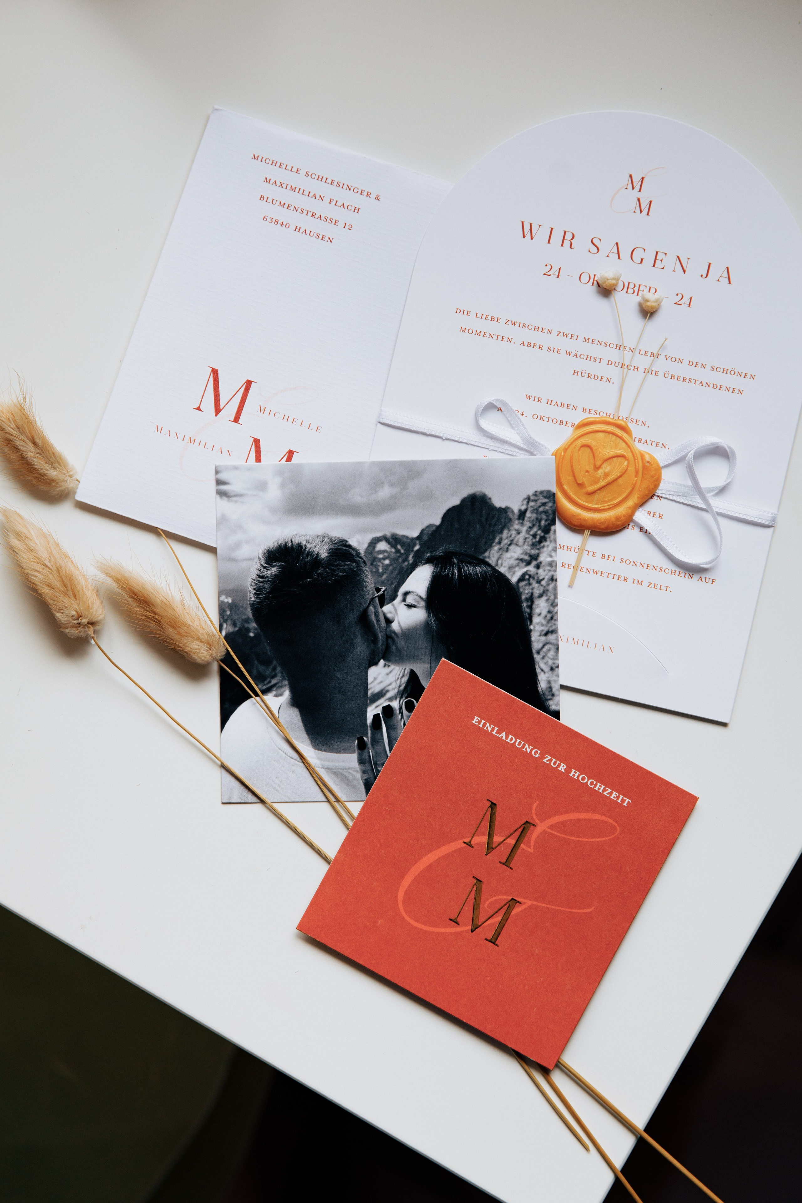 Stilvolle Hochzeitseinladungen in Weiß und Terrakotta mit Wachssiegel, begleitet von einem Schwarz-Weiß-Foto des Brautpaares beim Kuss vor einer Bergkulisse.