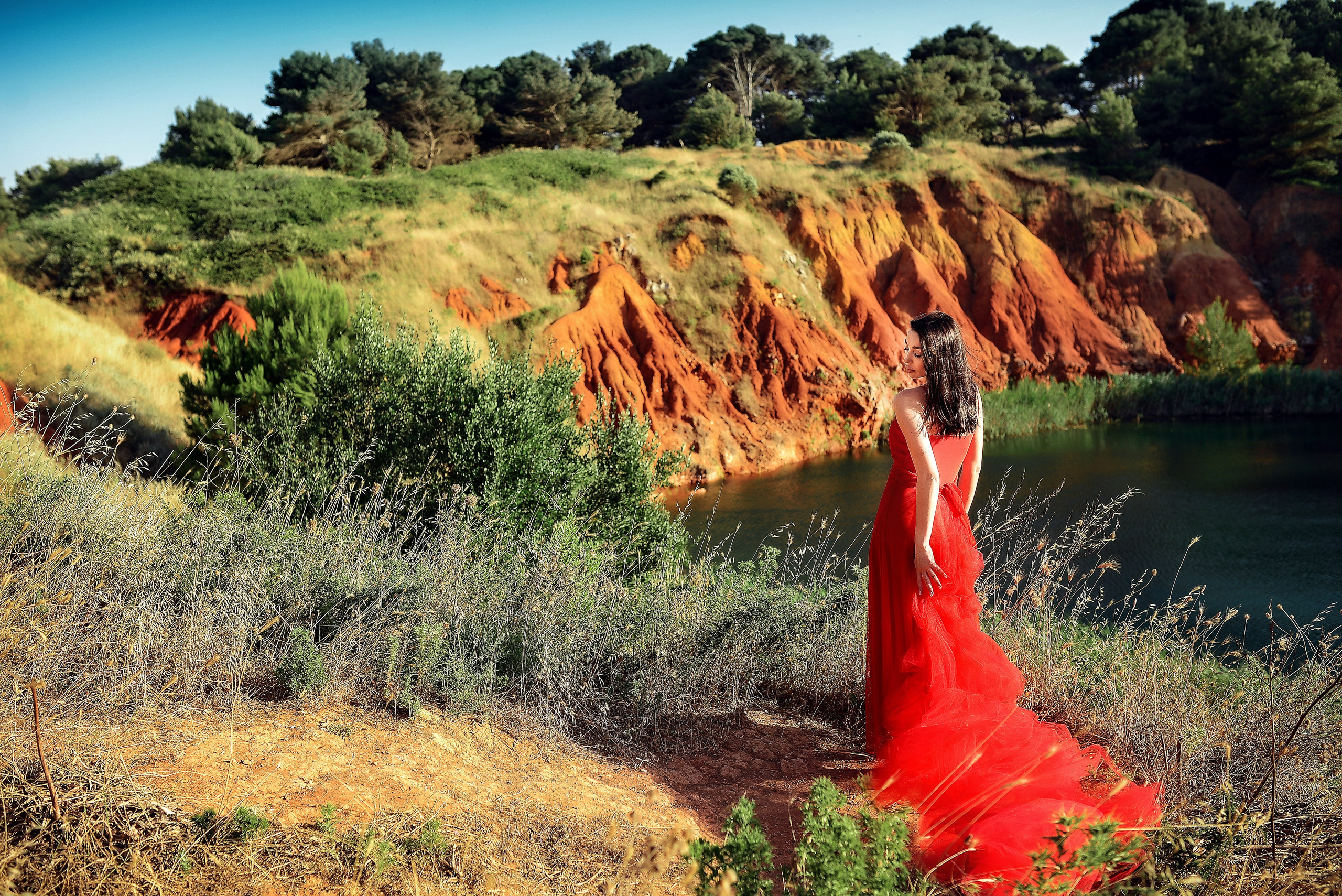 A woman in a red dress stands on the edge of a cliff by the Bauxite Quarry, Otranto in Salento, admiring the vibrant orange hills and lush greenery.