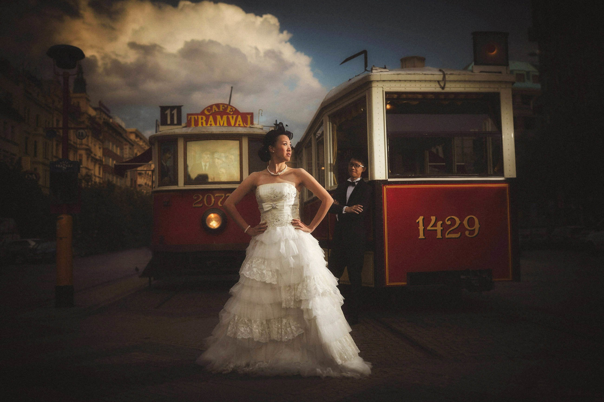 Confident Asian bride wearing a bird-cage veil poses stylishly in her wedding dress as her tuxedo-wearing groom stands behind her near two historical trams in Prague