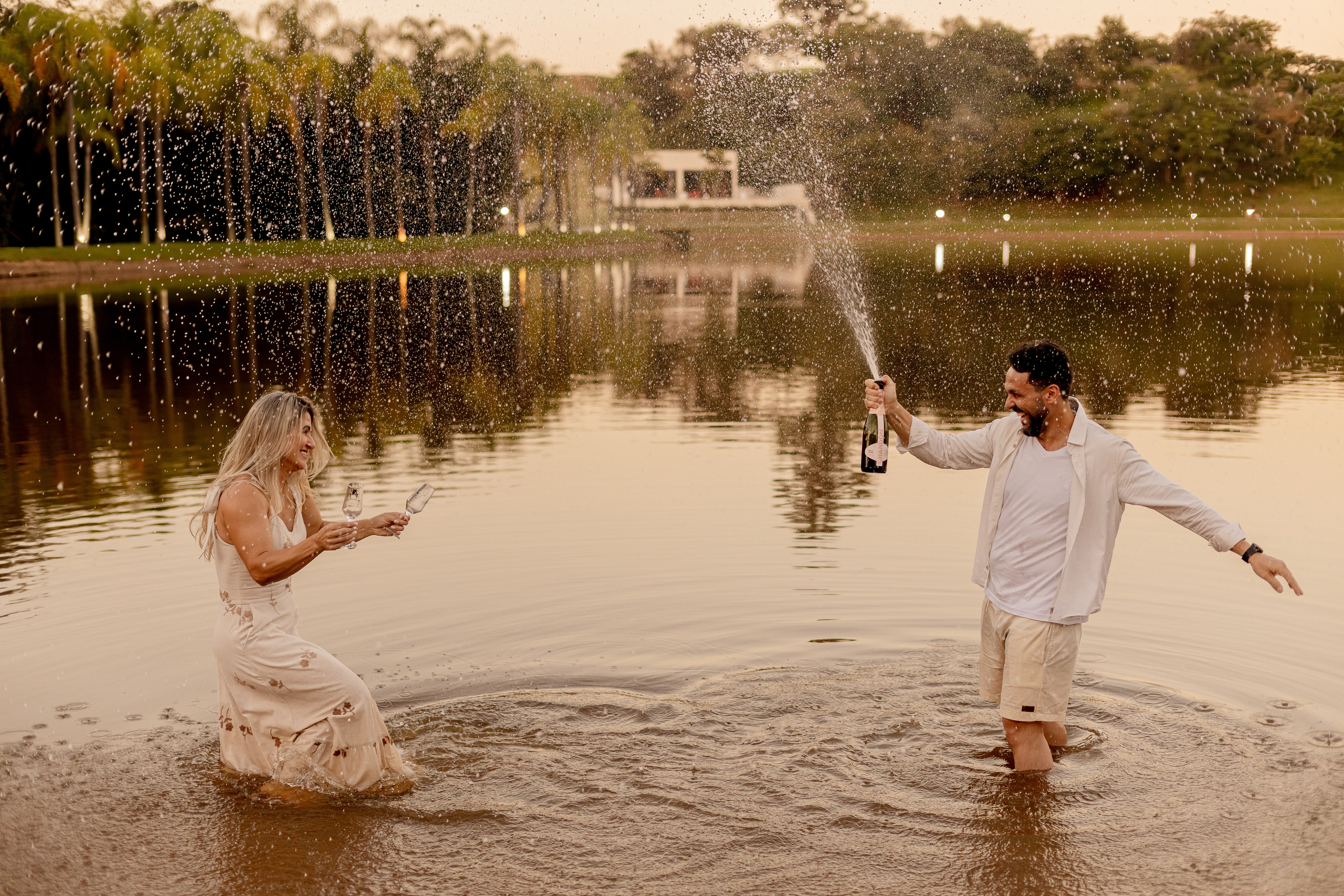Casal celebrando com champanhe dentro do lago no Resort das Oliveiras ao entardecer, com espirros d'água e sorrisos