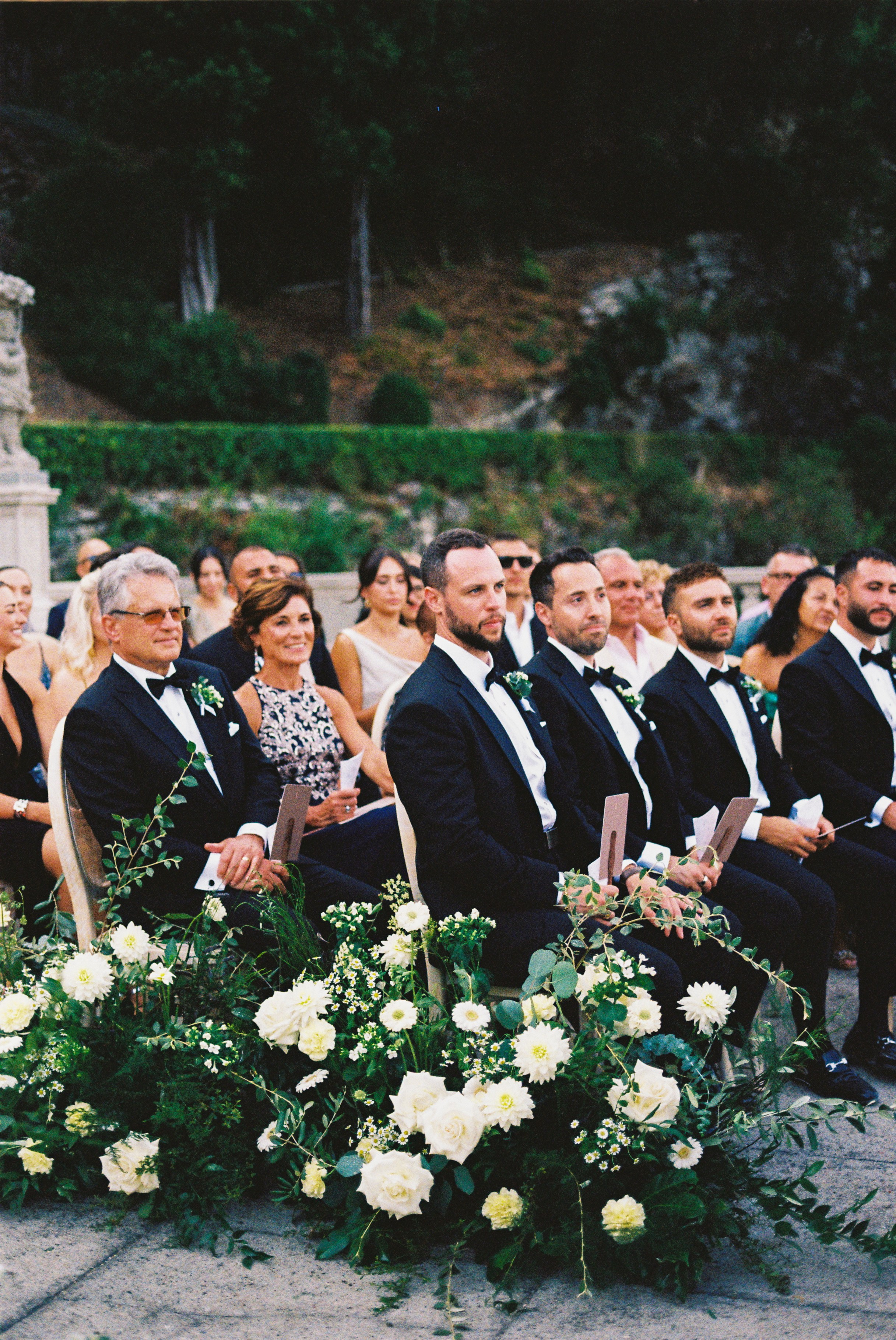 Groomsmen and family seated in formal attire with white floral arrangements at wedding ceremony.