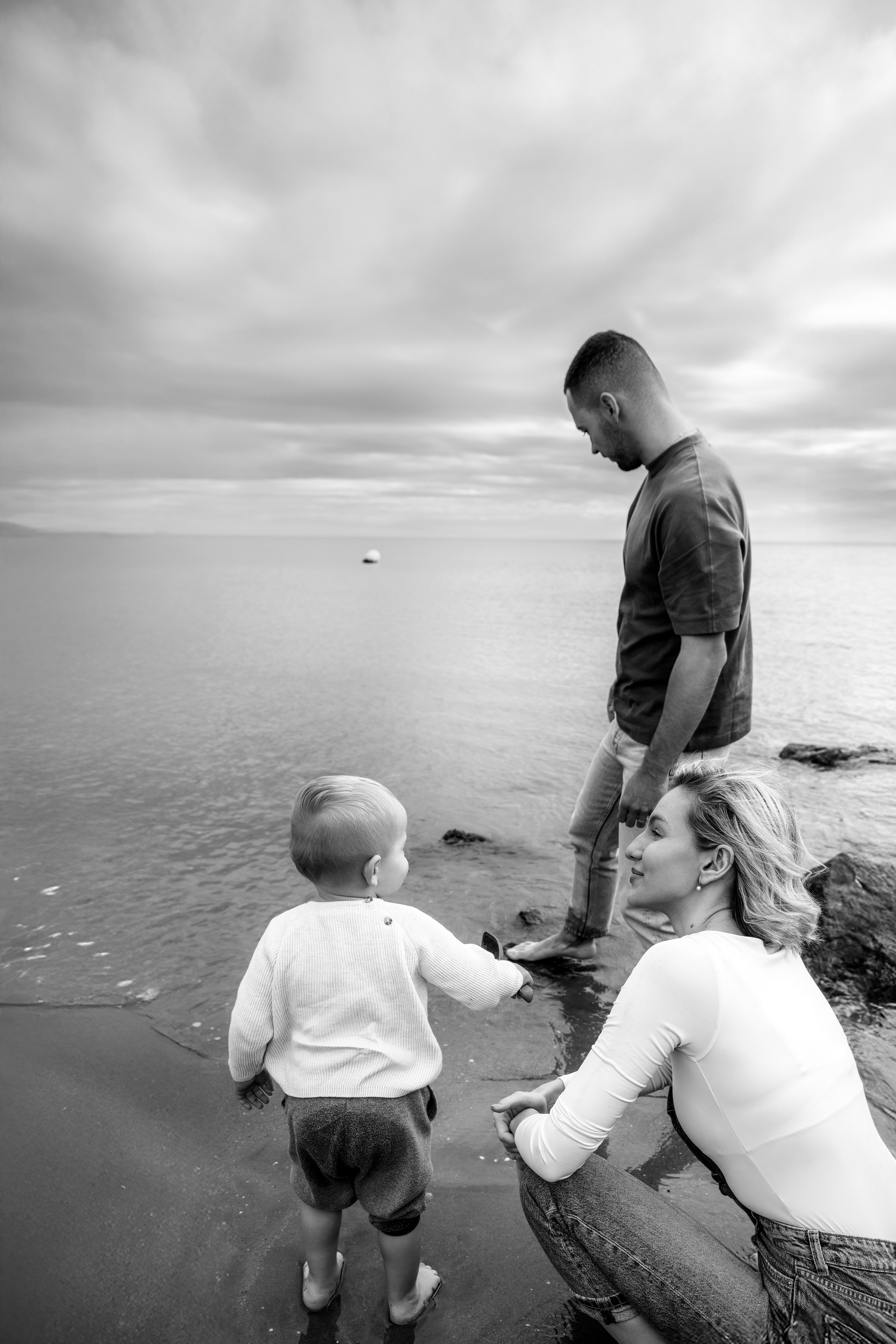 Sesión de fotos familiar en blanco y negro en la playa de Valencia, España — momento espontáneo de una joven familia disfrutando del mar, con un niño pequeño descalzo explorando la orilla. Ideal para quienes buscan sesiones familiares auténticas y emocionales en Valencia y zonas costeras de España.