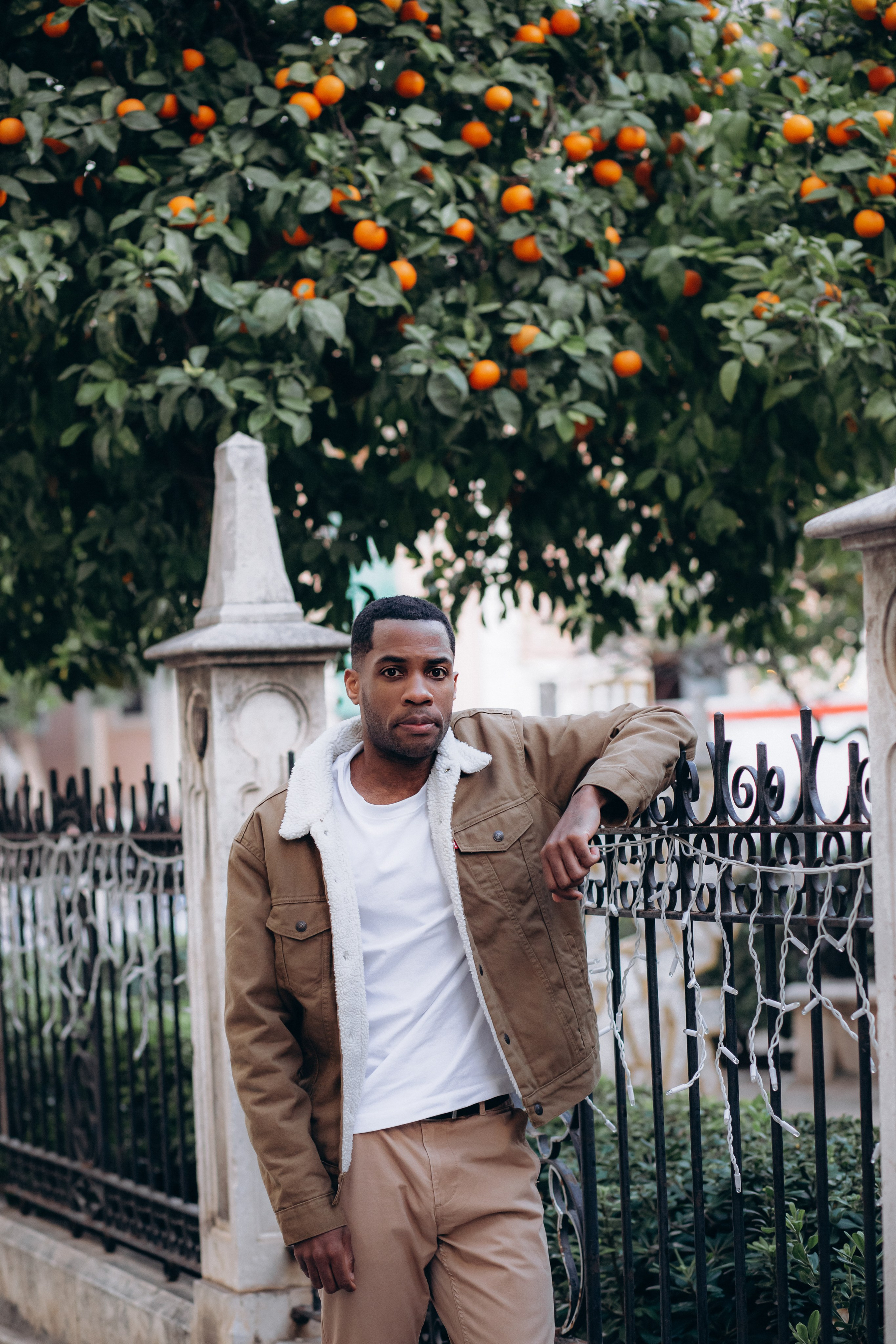 Мужская портретная фотосессия в Валенсии, Испания, с уверенным мужчиной, позирующим у кованого забора на фоне сада с апельсиновыми деревьями, в повседневной куртке и белой рубашке, снятым при естественном дневном свете с расслабленной средиземноморской атмосферой — идеально для мужской портретной фотографии, lifestyle-фотосессий для мужчин, портретов для персонального бренда, городских outdoor-портретов и профессиональных мужских фотосессий в Валенсии и по всей Испании.