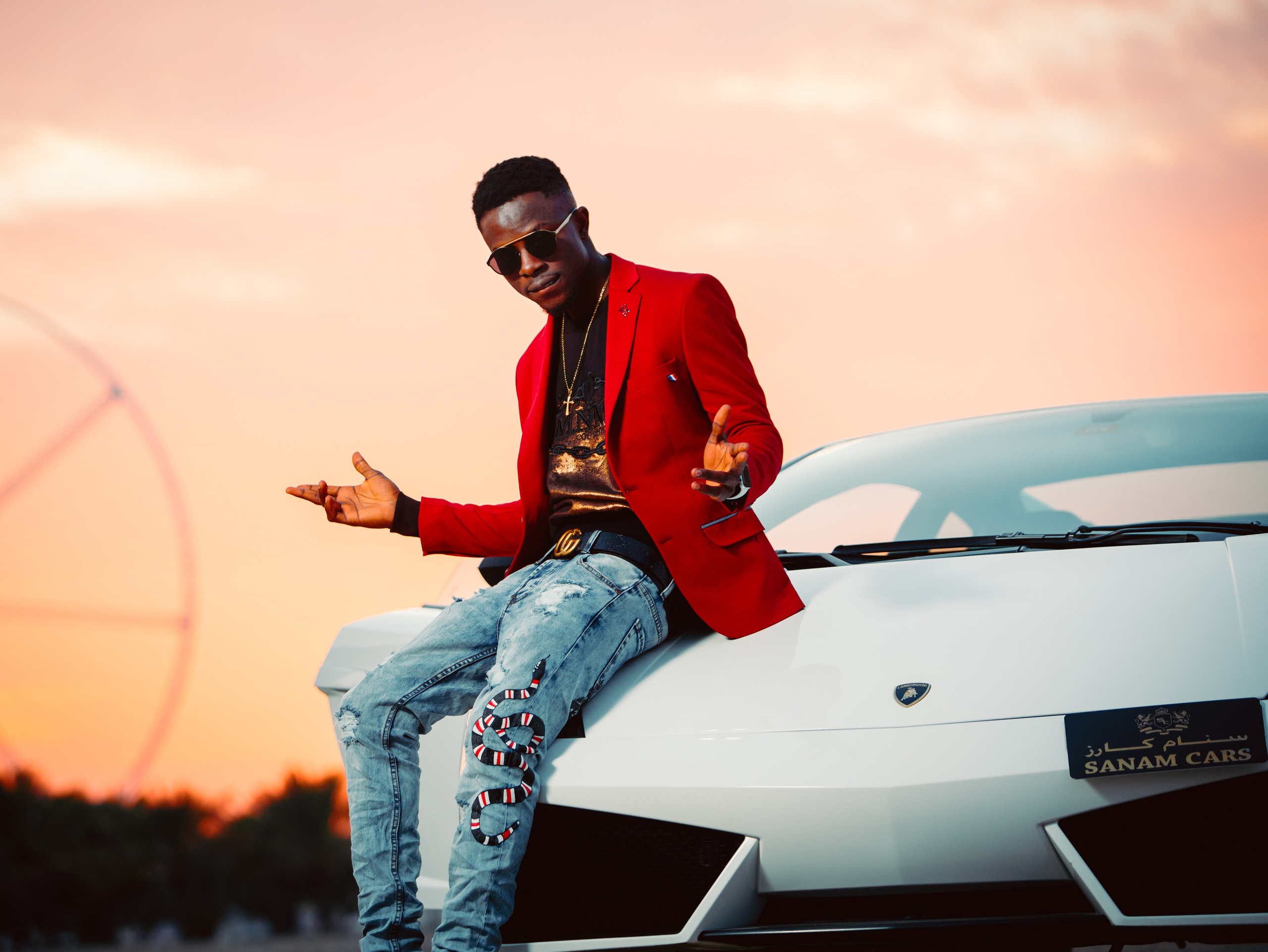A man sits on a white Ferrari, wearing a red blazer, against the backdrop of a Dubai sunset