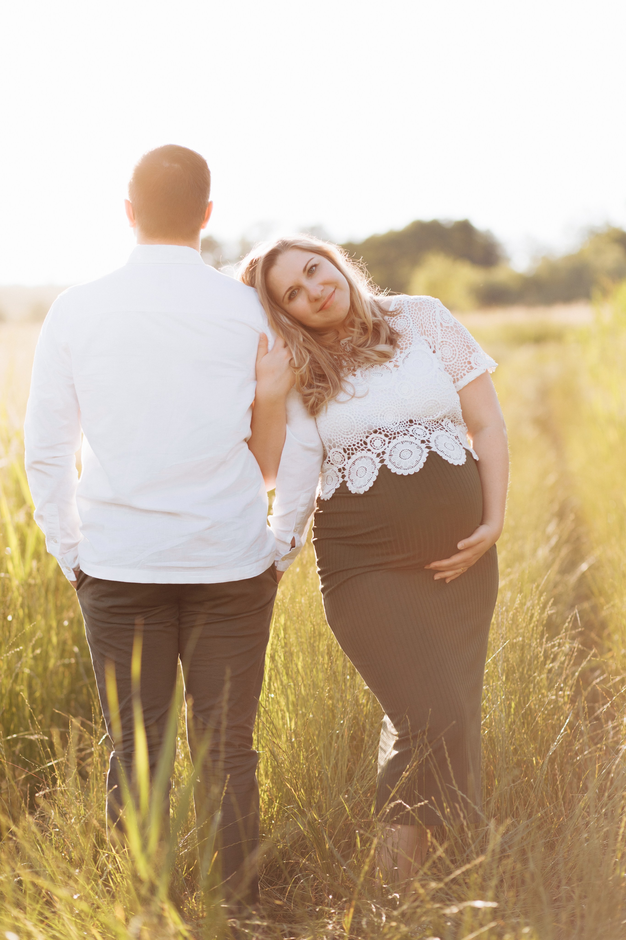 Frau hält ihren Babybauch und lehnt sich im Feld während eines Fotoshootings an die Schulter ihres Mannes.