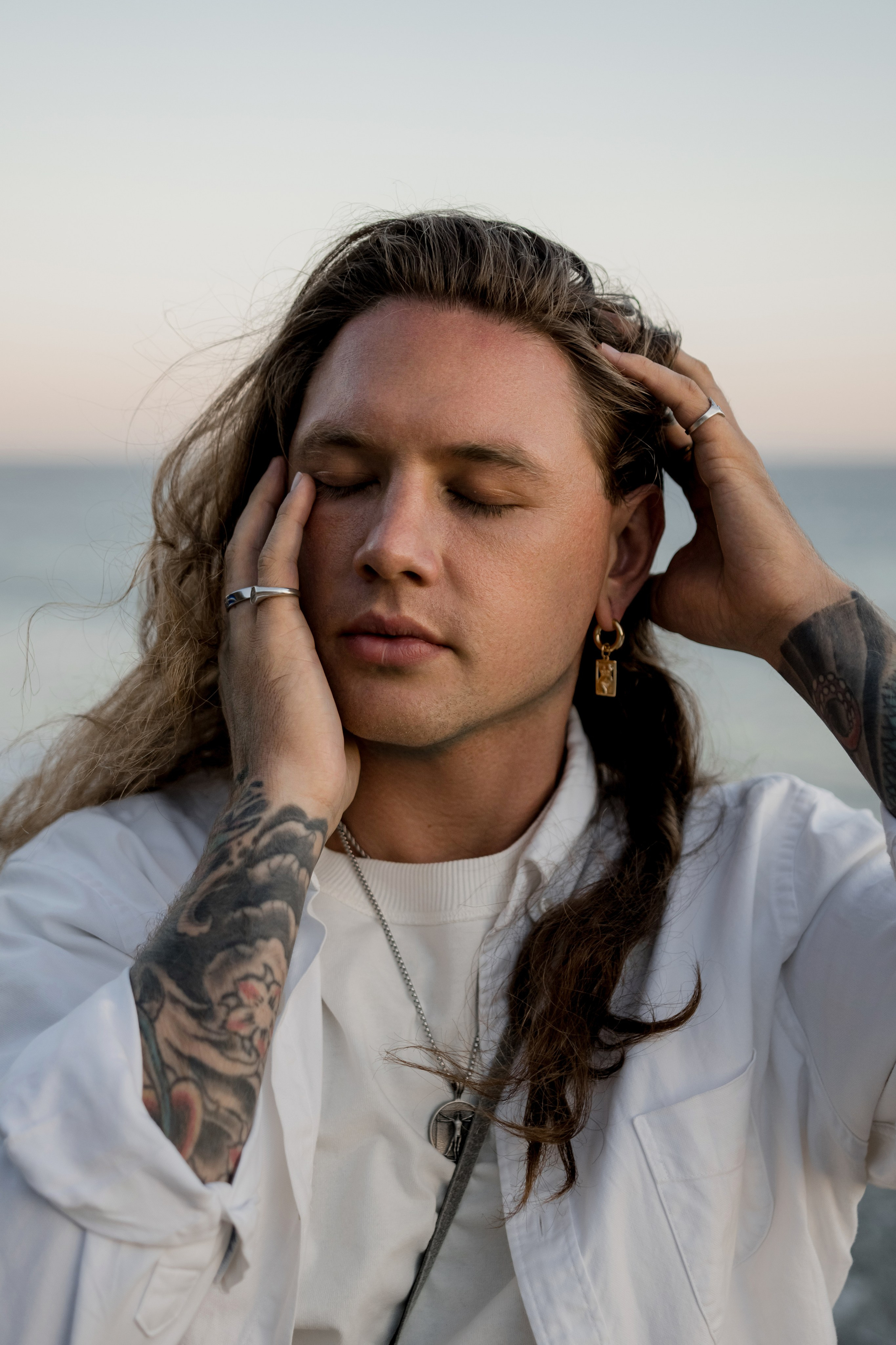 Male model with long hair and tattoos posing by the ocean, emphasizing relaxed and confident style.