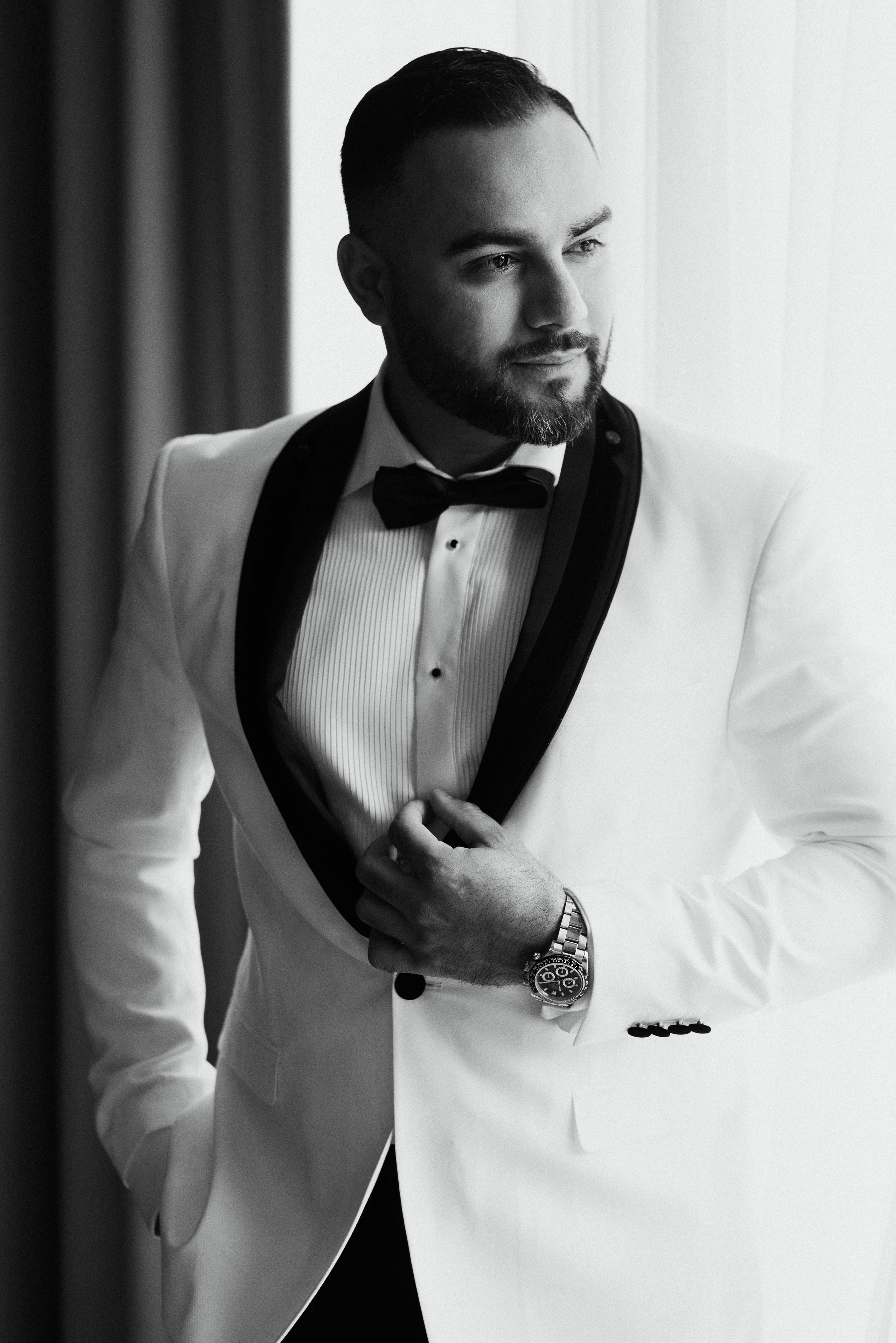 Black and white portrait of a groom in a stylish white tuxedo with black lapels, preparing for his wedding day