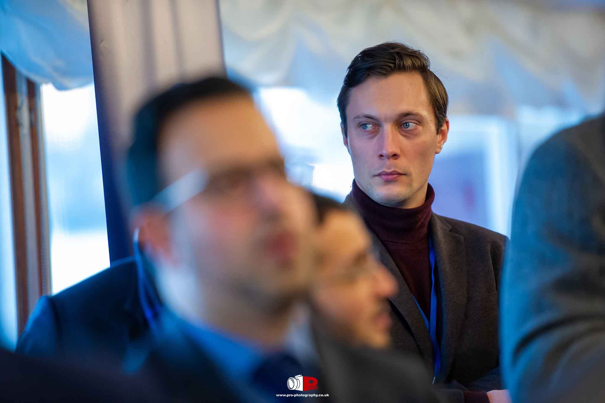 A young businessman in a dark blazer and maroon turtleneck attentively observing the surroundings at a professional networking event.