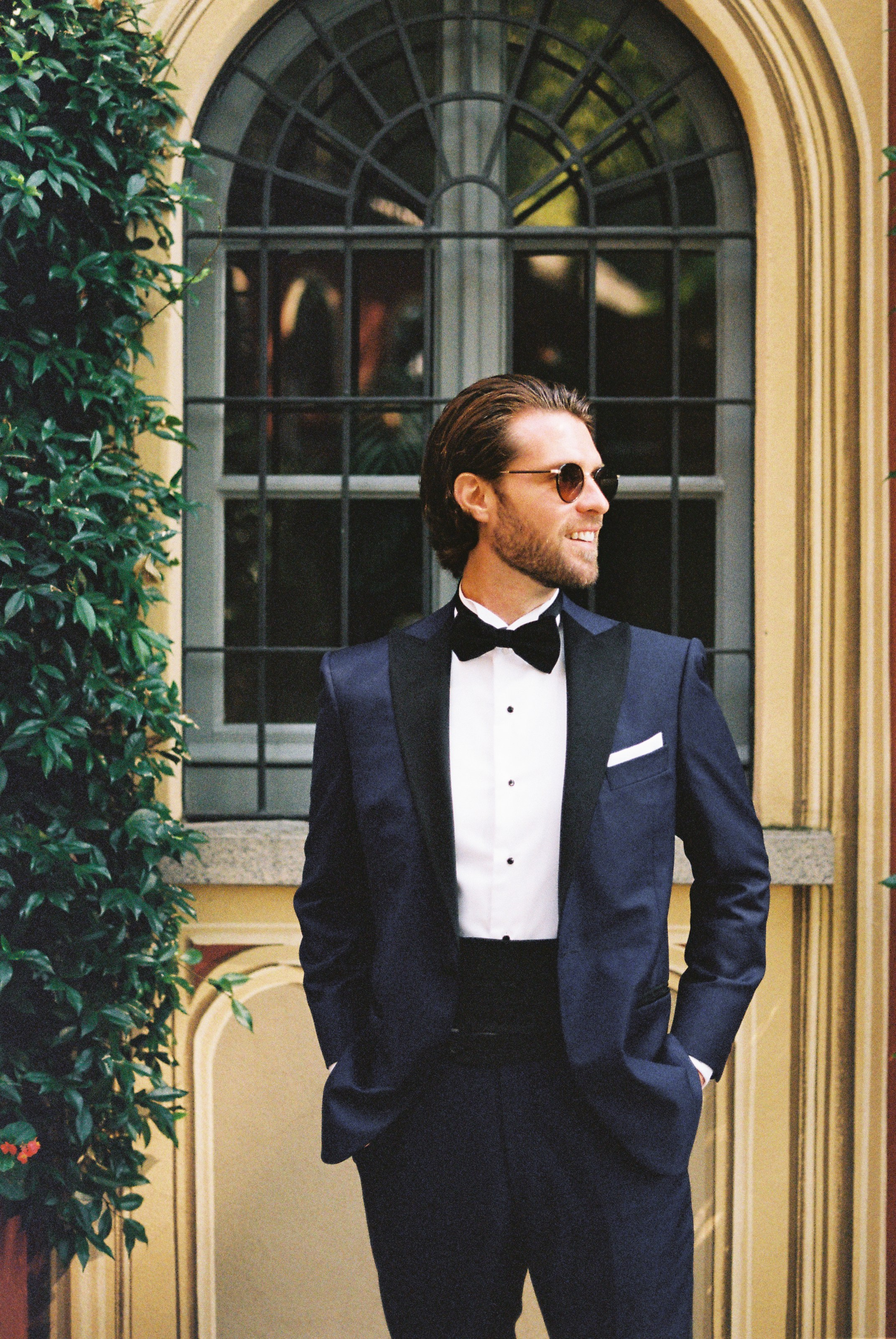 Groom in a stylish blue tuxedo stands confidently against an elegant arched window backdrop.
