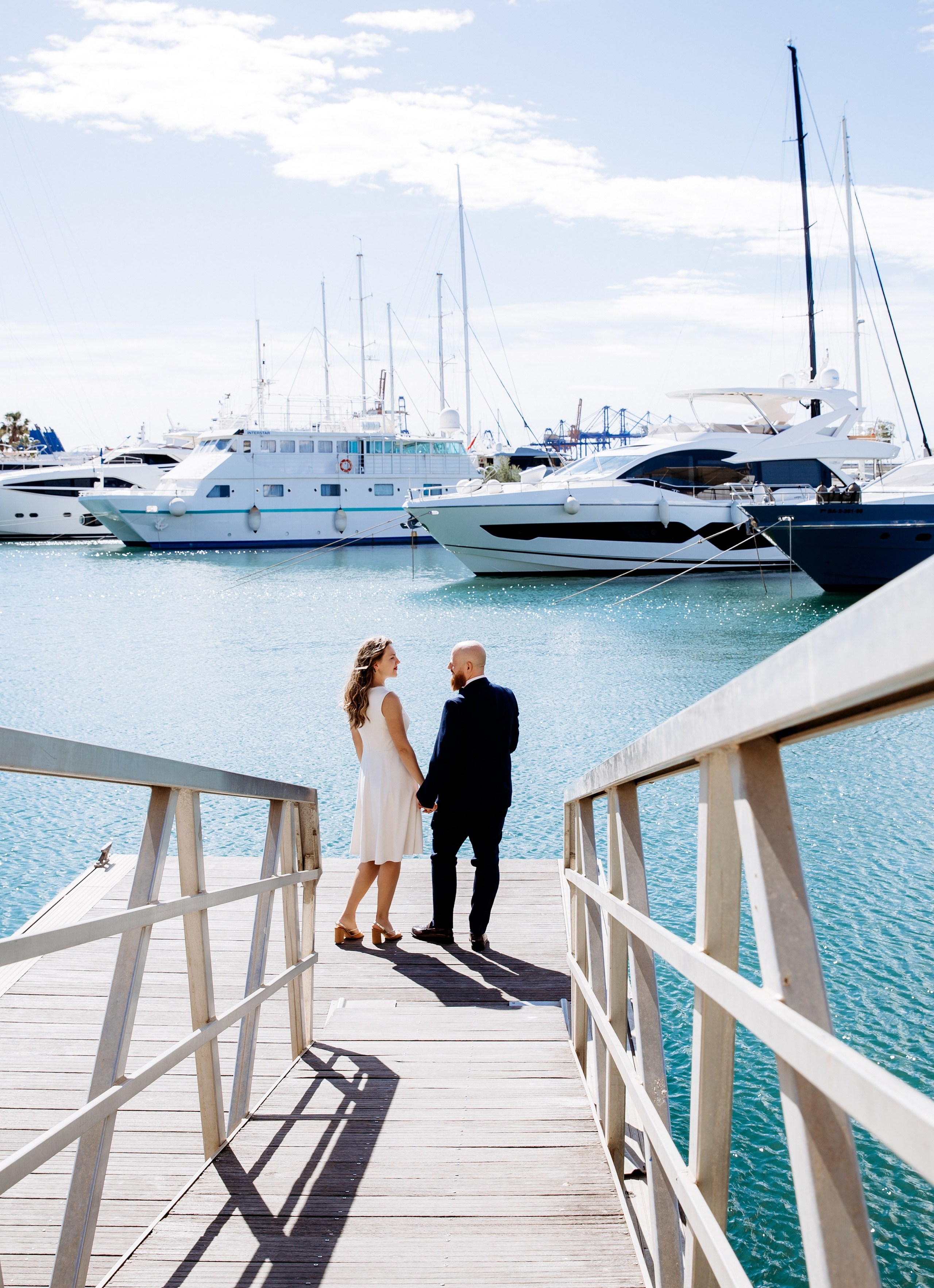 Bride and groom walking hand in hand on a marina pier in València, Spain, with luxury yachts in the background, captured in a natural destination wedding photography style.