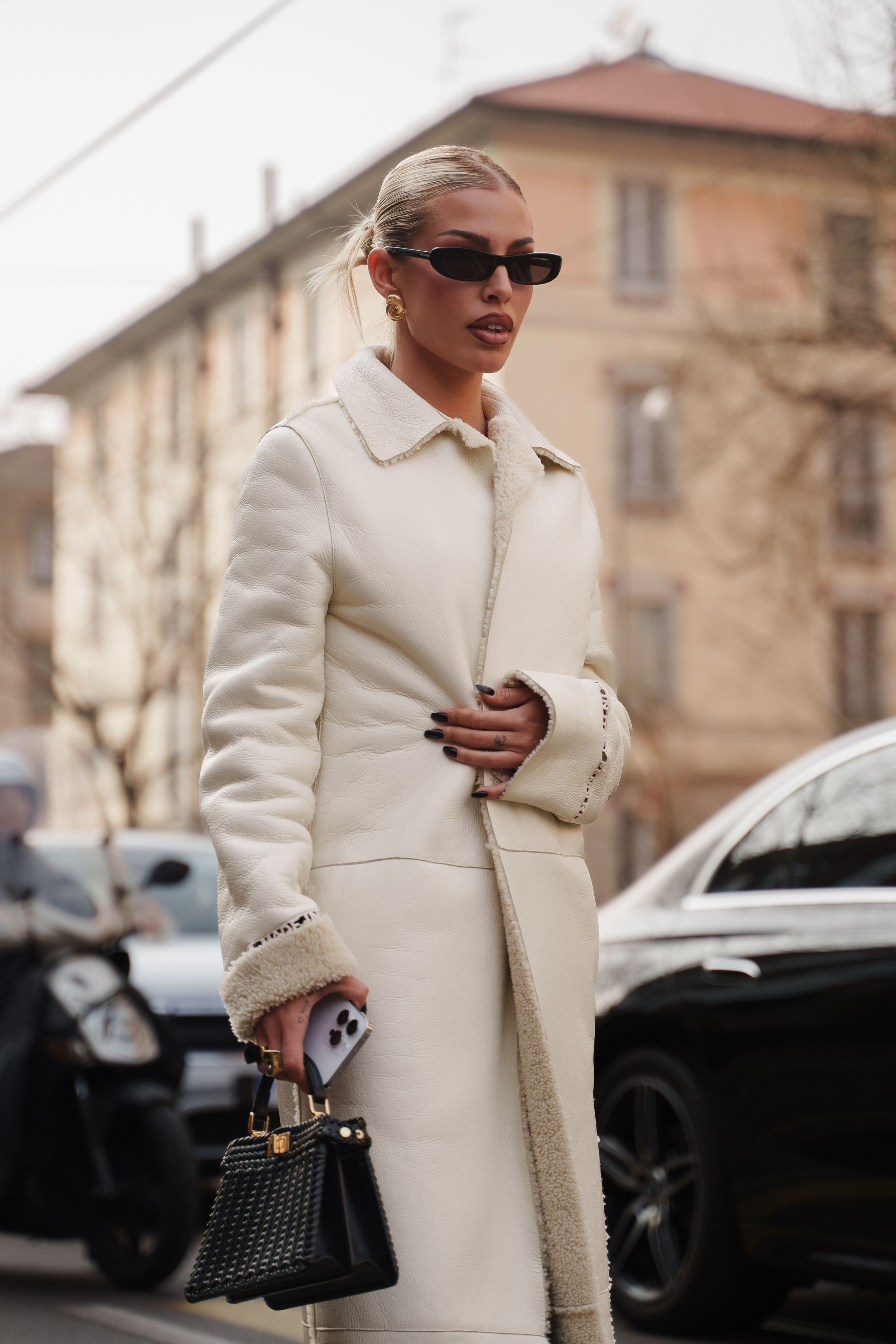 Blonde woman in a white shearling coat holding a small black handbag, gazing to the side
