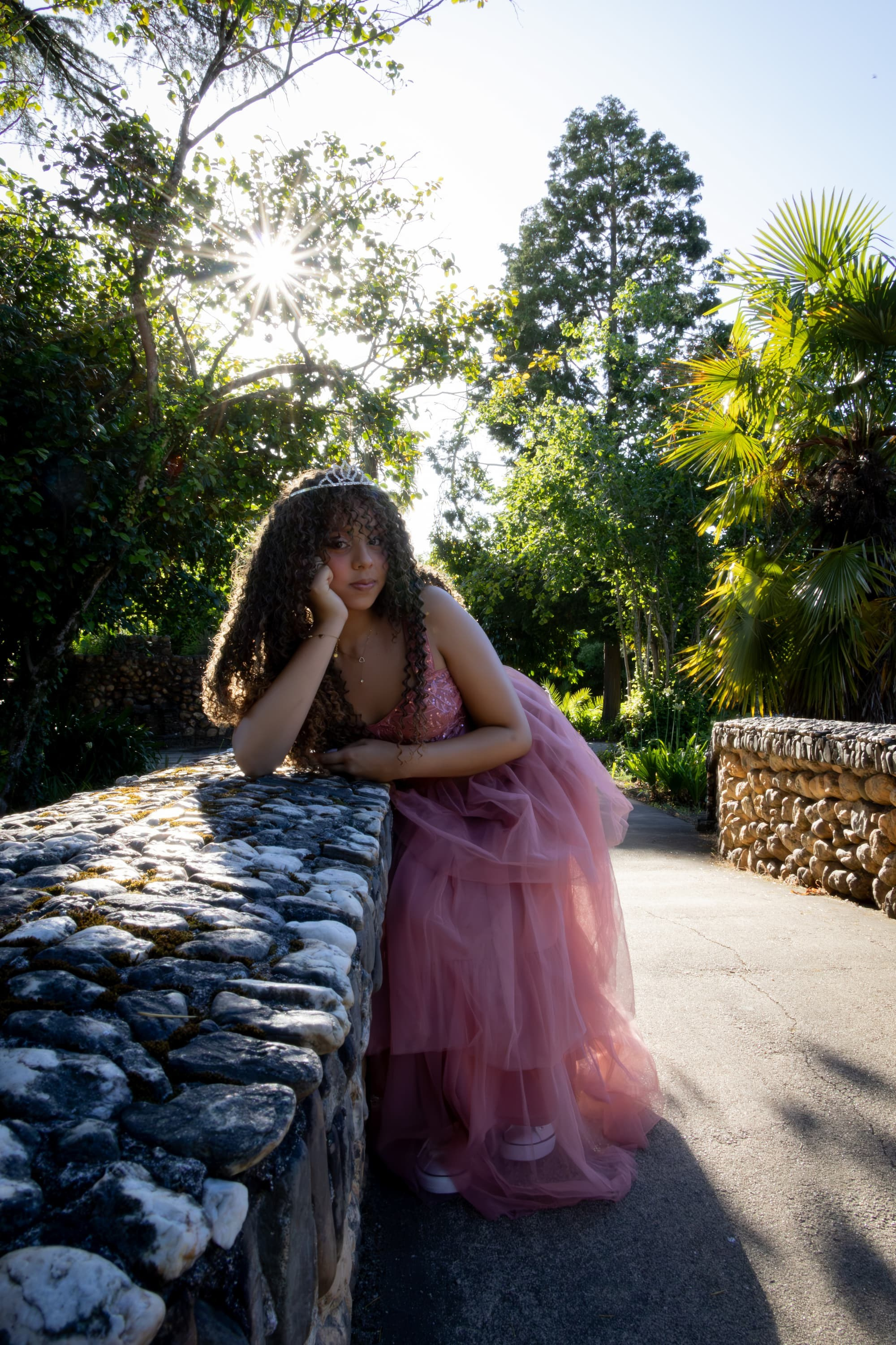 Uma jovem em pose pensativa sobre um muro de pedras rústicas, usando um vestido volumoso rosa de tule, tiara e tênis brancos. Cenário de jardim botânico verdejante com luz do sol forte.
