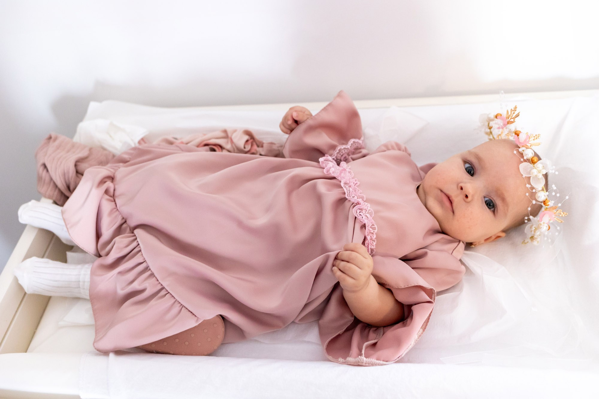 Bebelus îmbrăcat într-o rochiță roz elegantă cu tulle, așezat pe un pat în timpul pregătirilor de dimineață.
