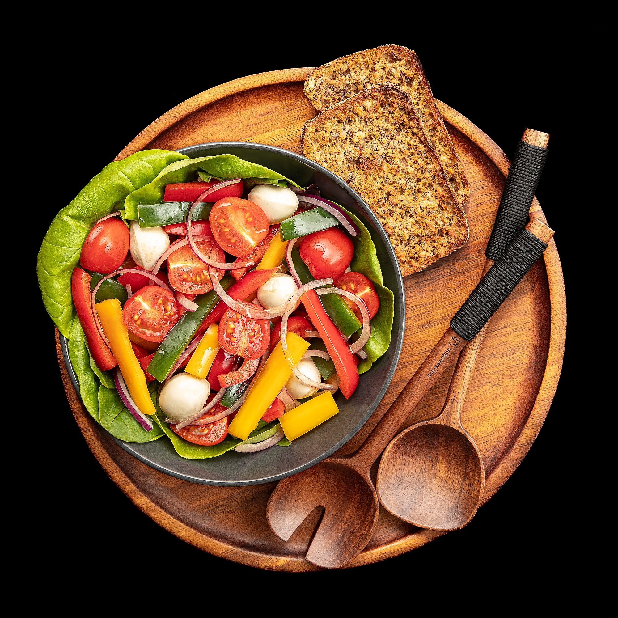 Photographer Roman Djuzev - Photo of vegetarian salad in Flat Lay style on a wooden platter with wooden spoons on a black background, salad of sweet peppers, tomato, herbs, red onion and cheese in a black plate.