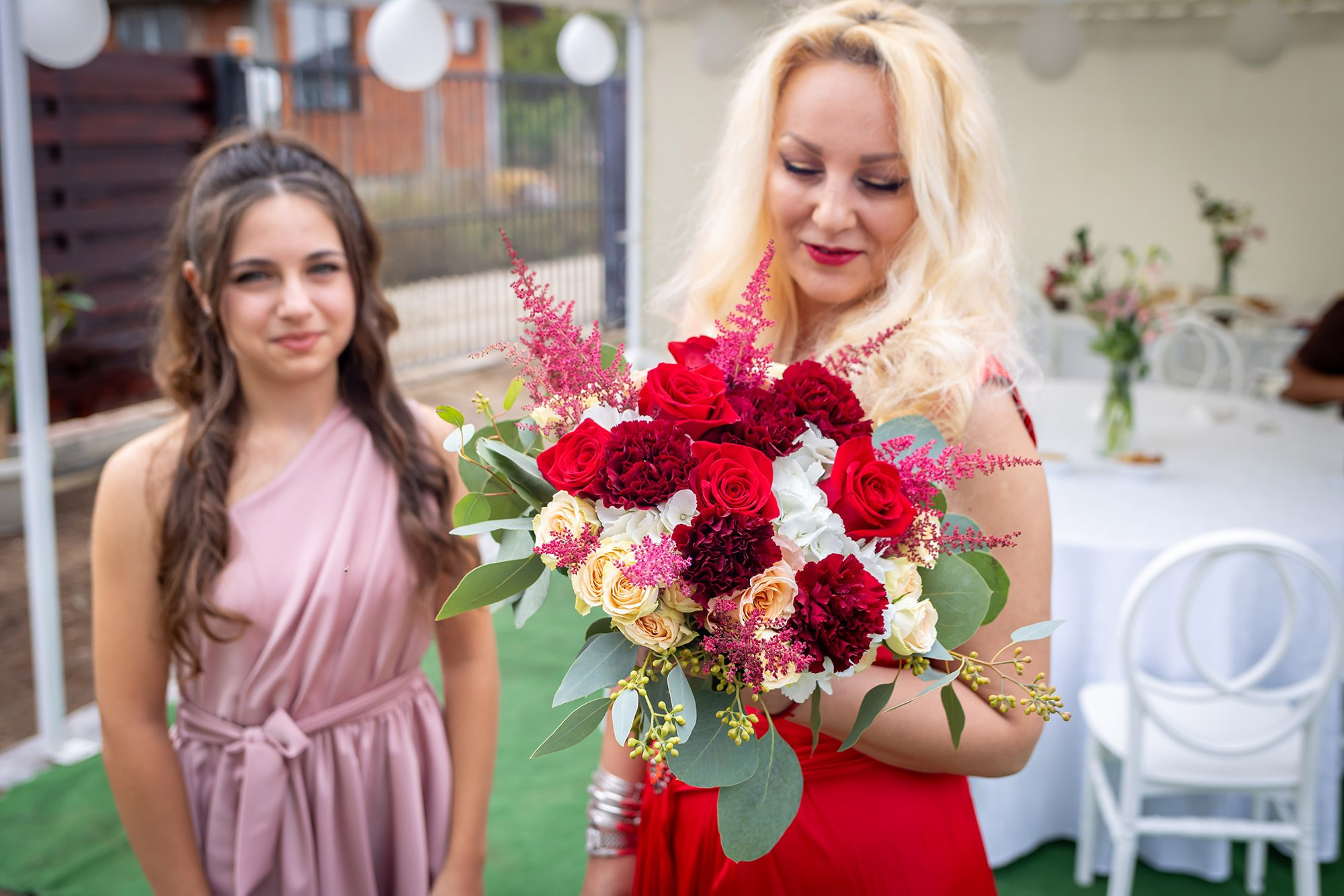 Nașa în rochie roșie ținând un buchet de mireasă cu trandafiri roșii și bujori, alături de o tânără în rochie roz