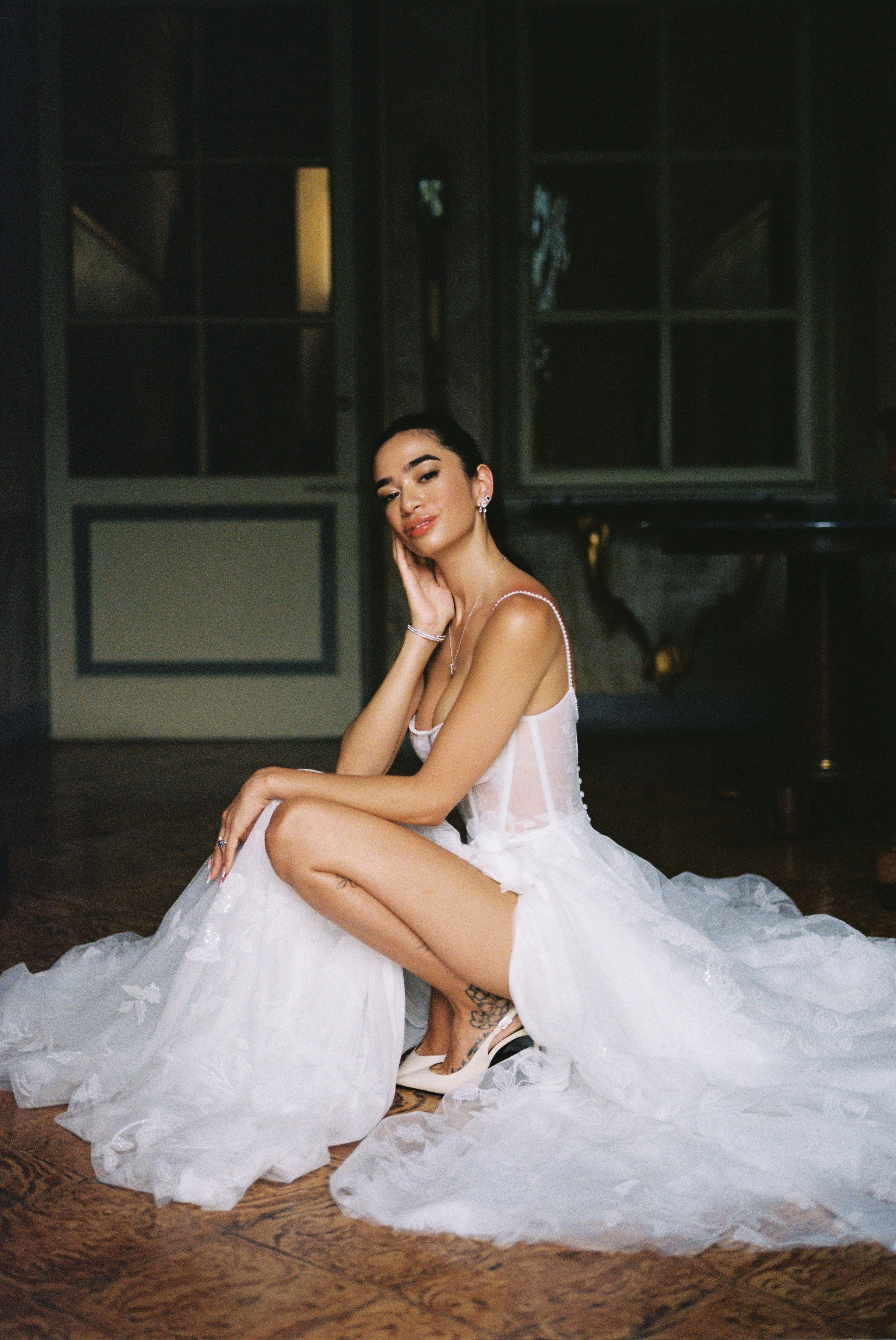 Bride in delicate white gown seated on floor, posing thoughtfully in elegant, dim room.