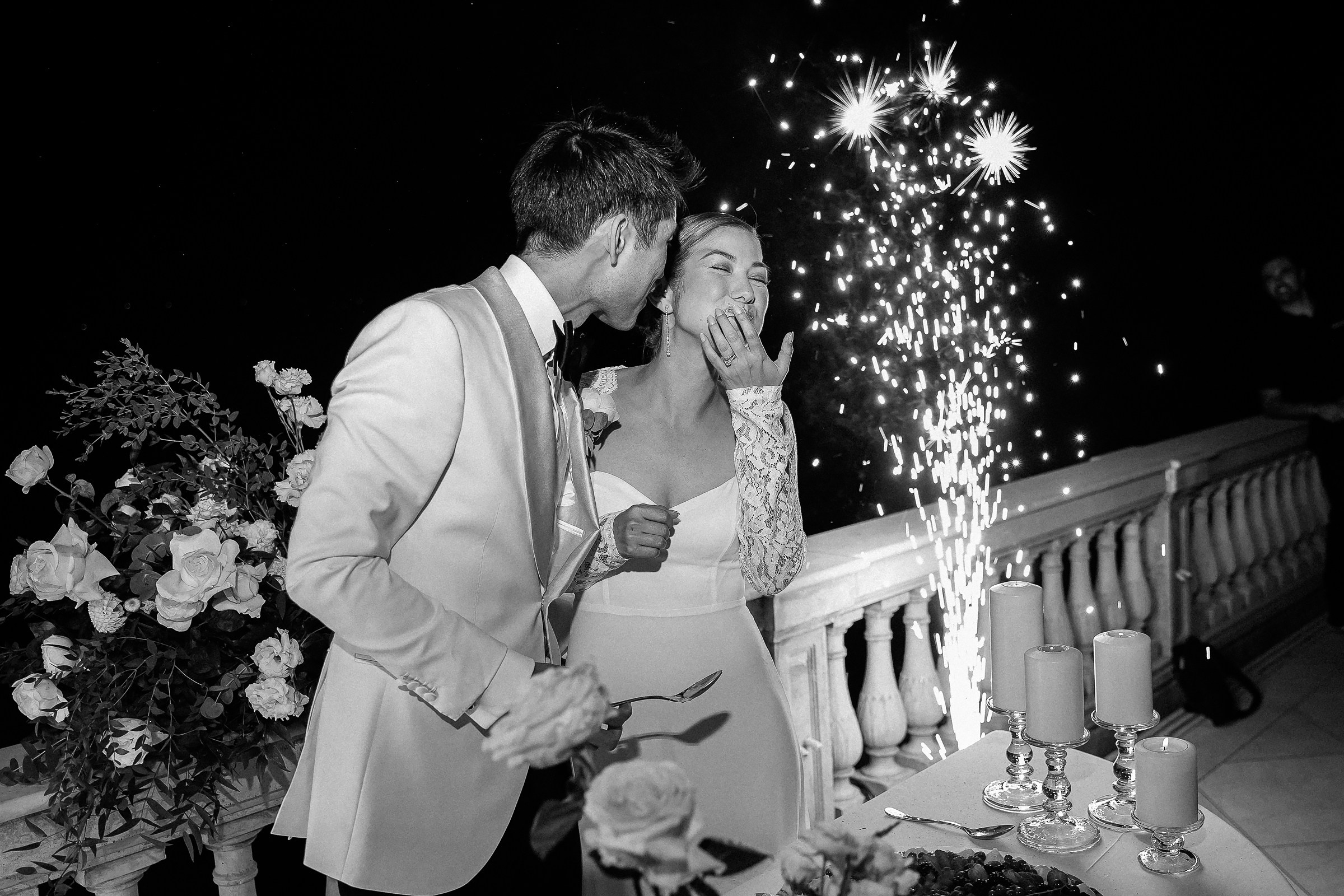 Groom playfully smears cake on bride’s face with sparklers illuminating the background.
