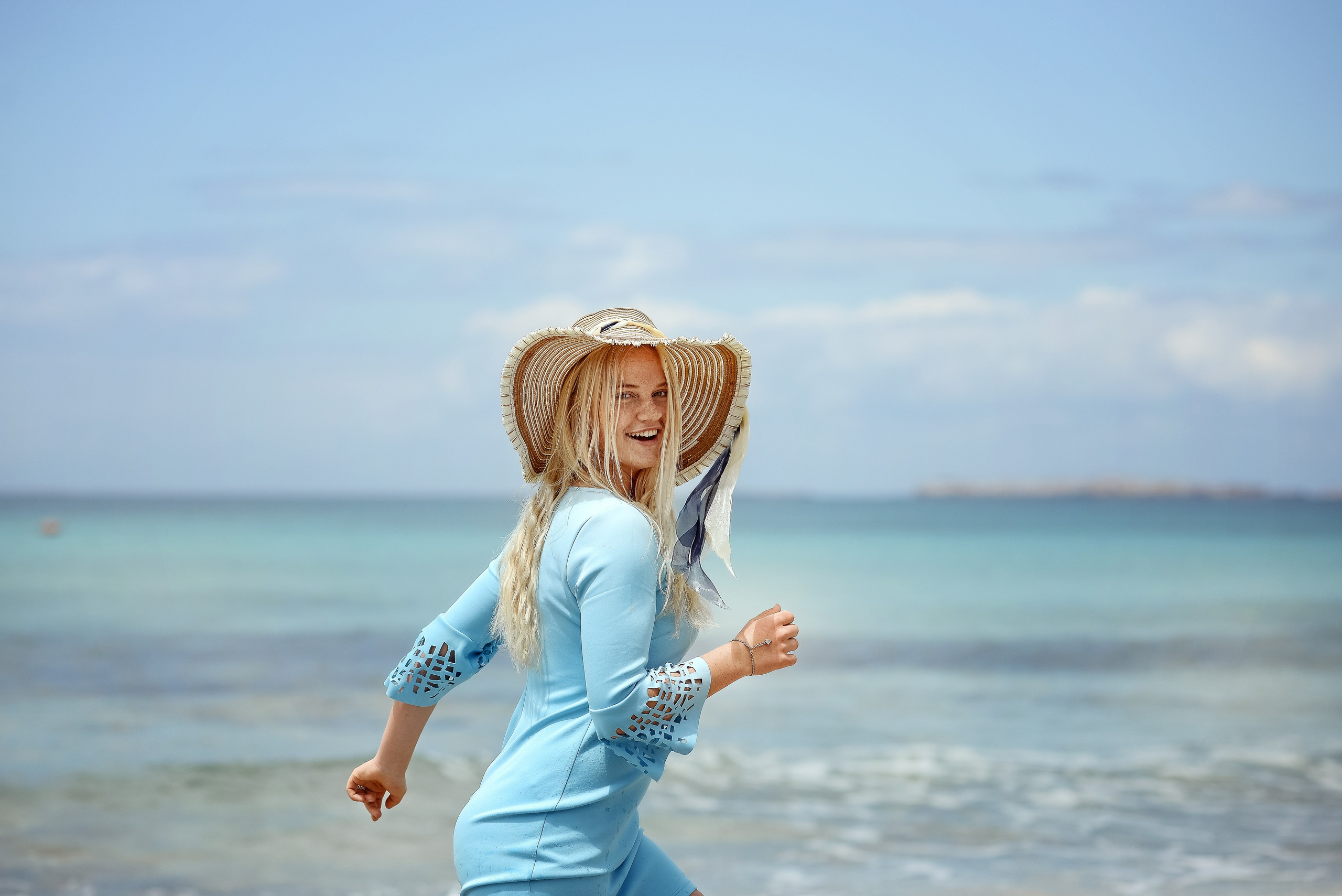 A smiling blonde woman in a light blue dress and straw hat runs along the seashore in Salento. The turquoise water and clear sky evoke a sense of freedom and joy.