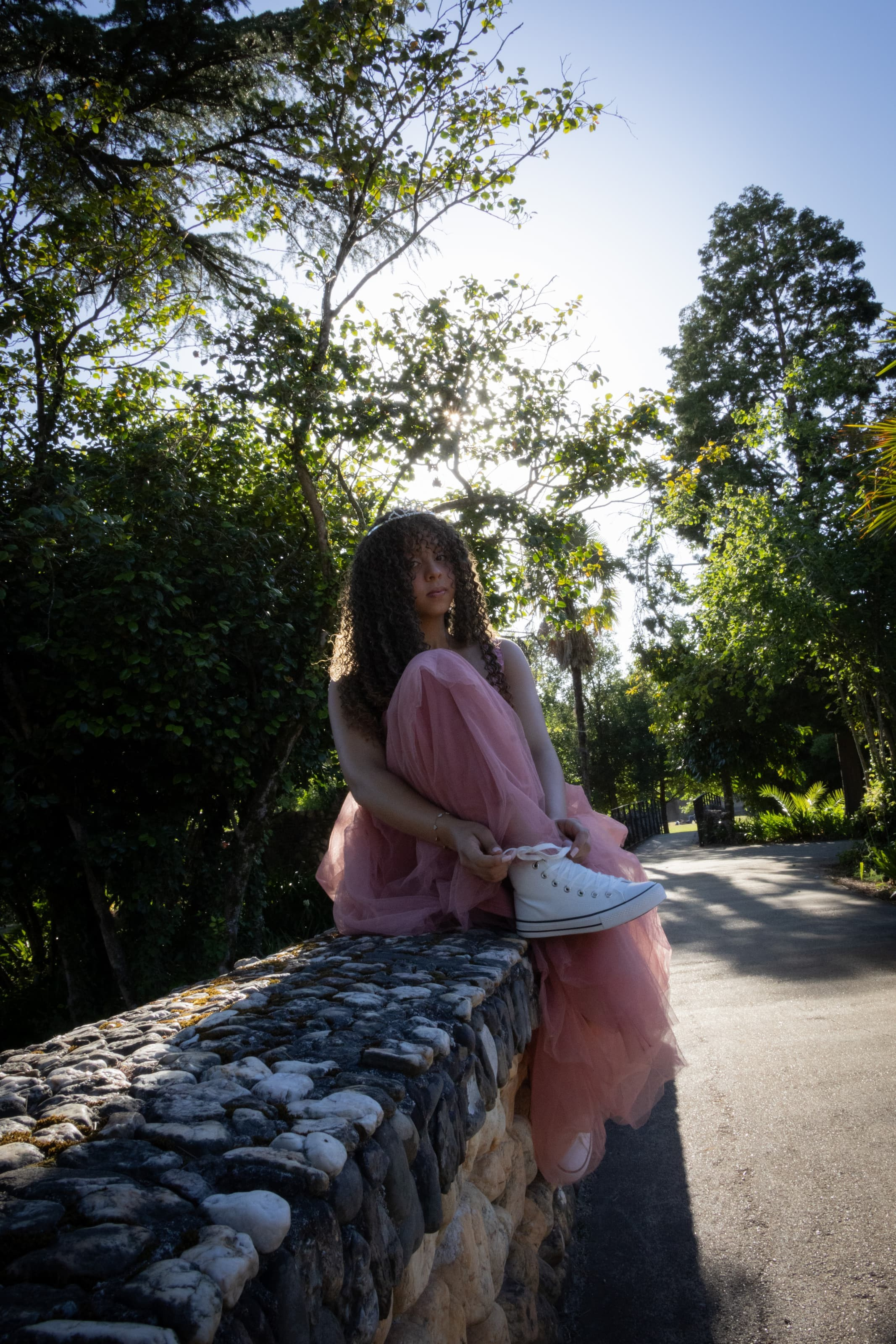 Jovem de vestido rosa longo sentada no parapeito de uma ponte de pedra antiga dentro de um bosque com luz solar filtrada pelas folhas.