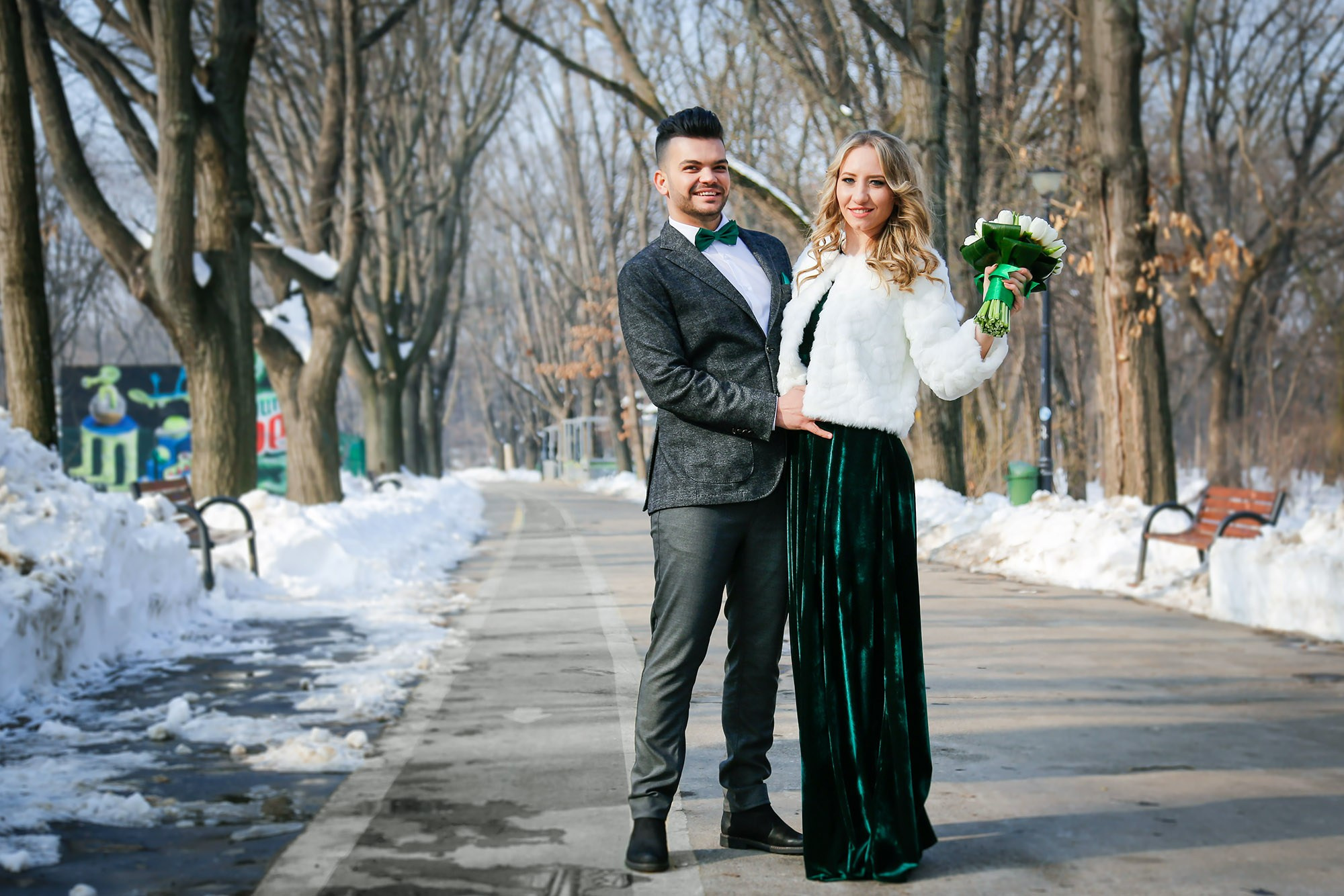 Wide shot of the newlyweds walking down a snowy park pathway, surrounded by trees completely covered in snow.