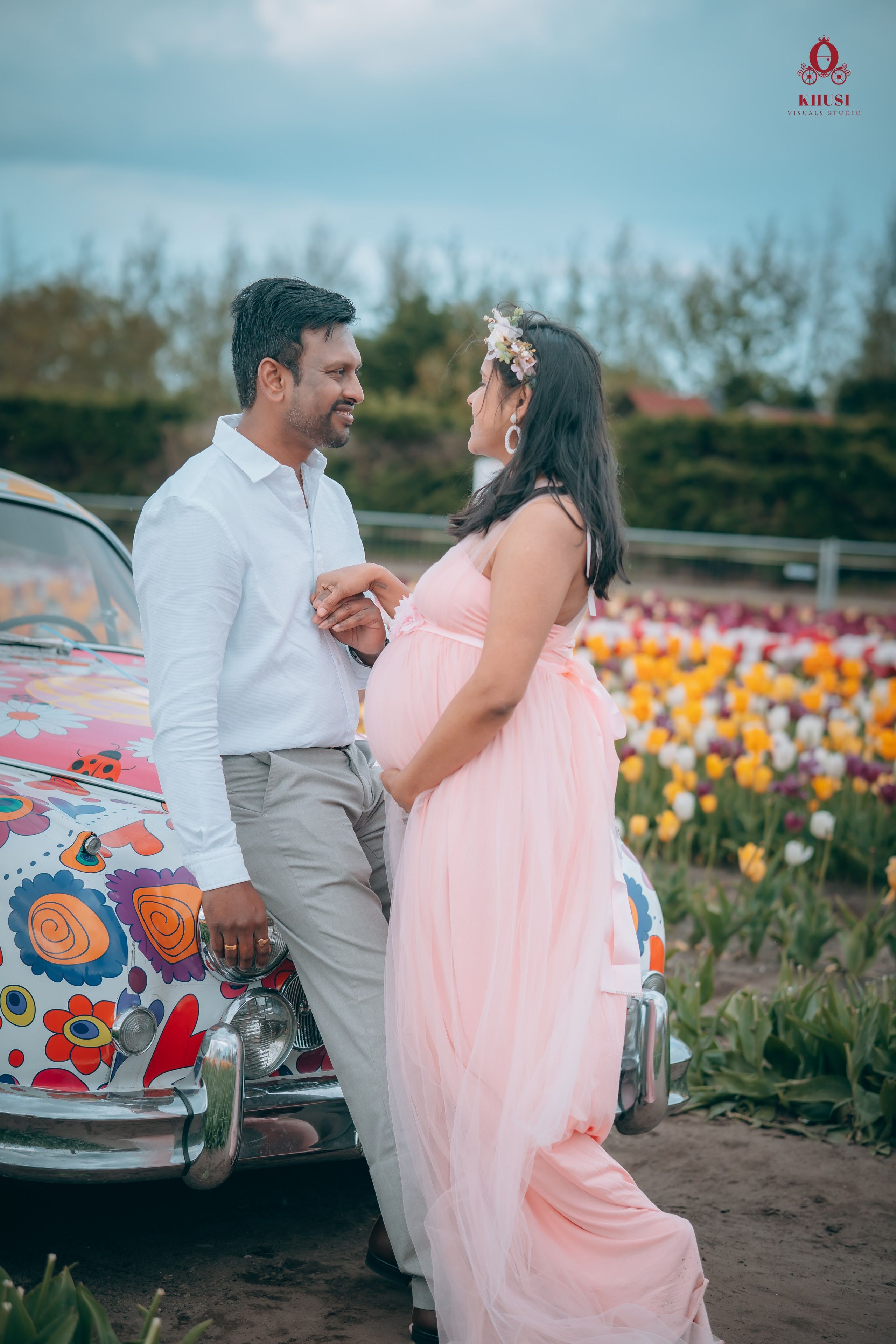 A pregnant woman holding hand of her husband and leaning on a car with floral print in a tulip field in Netherlands