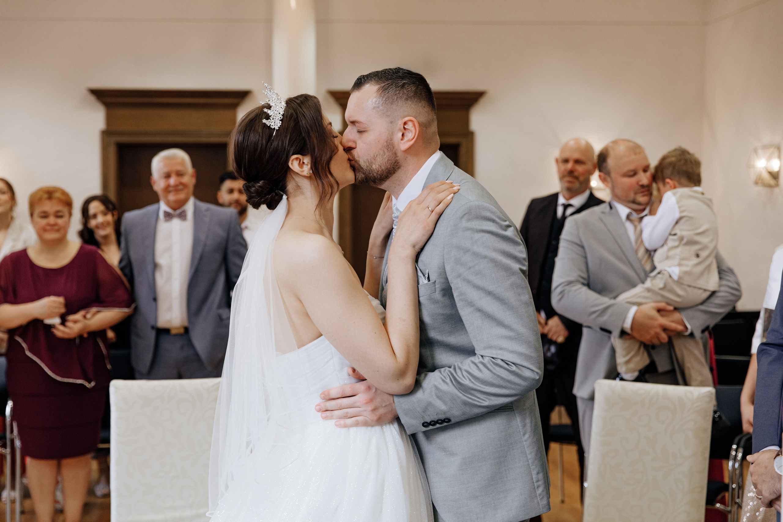Der erste Kuss des Brautpaares, während im Hintergrund die Gäste lächeln. Ein romantischer Moment der Hochzeit.