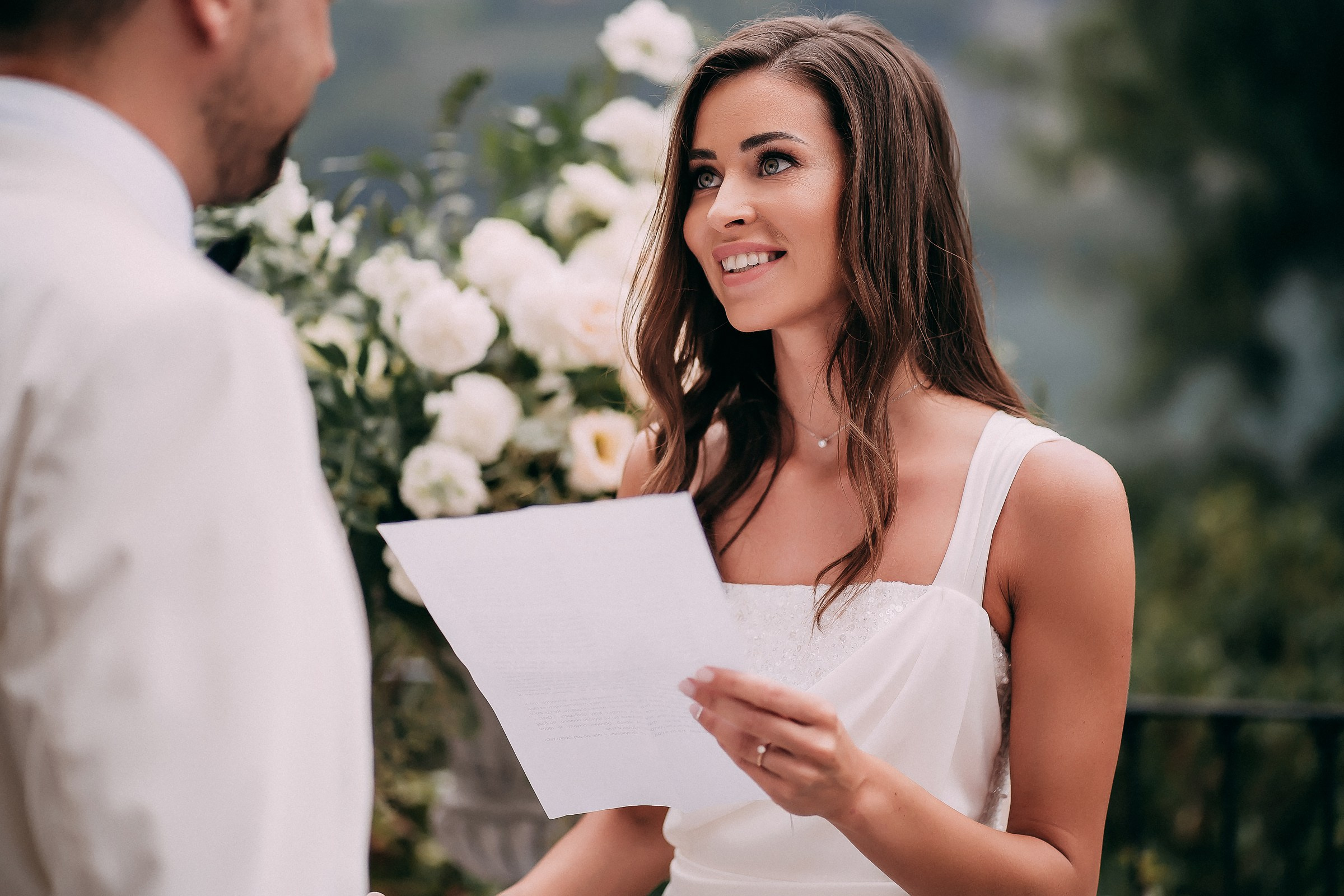 Bride in elegant white dress reading vows in front of lush floral backdrop at outdoor wedding ceremony. 
