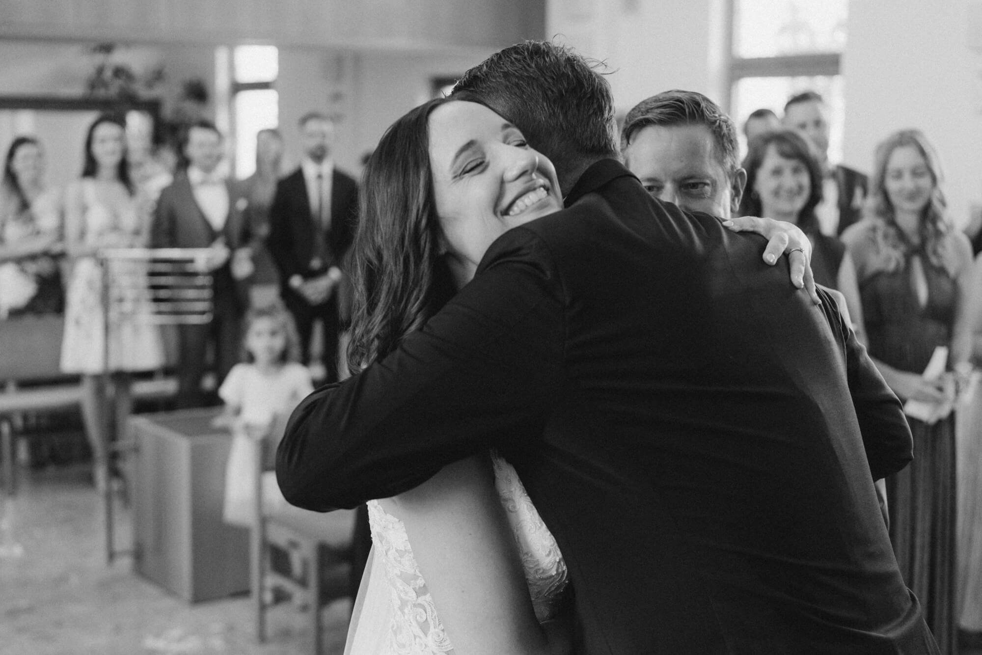 Bride laughing and embracing someone after the ceremony, guests applauding and smiling in the background; black and white