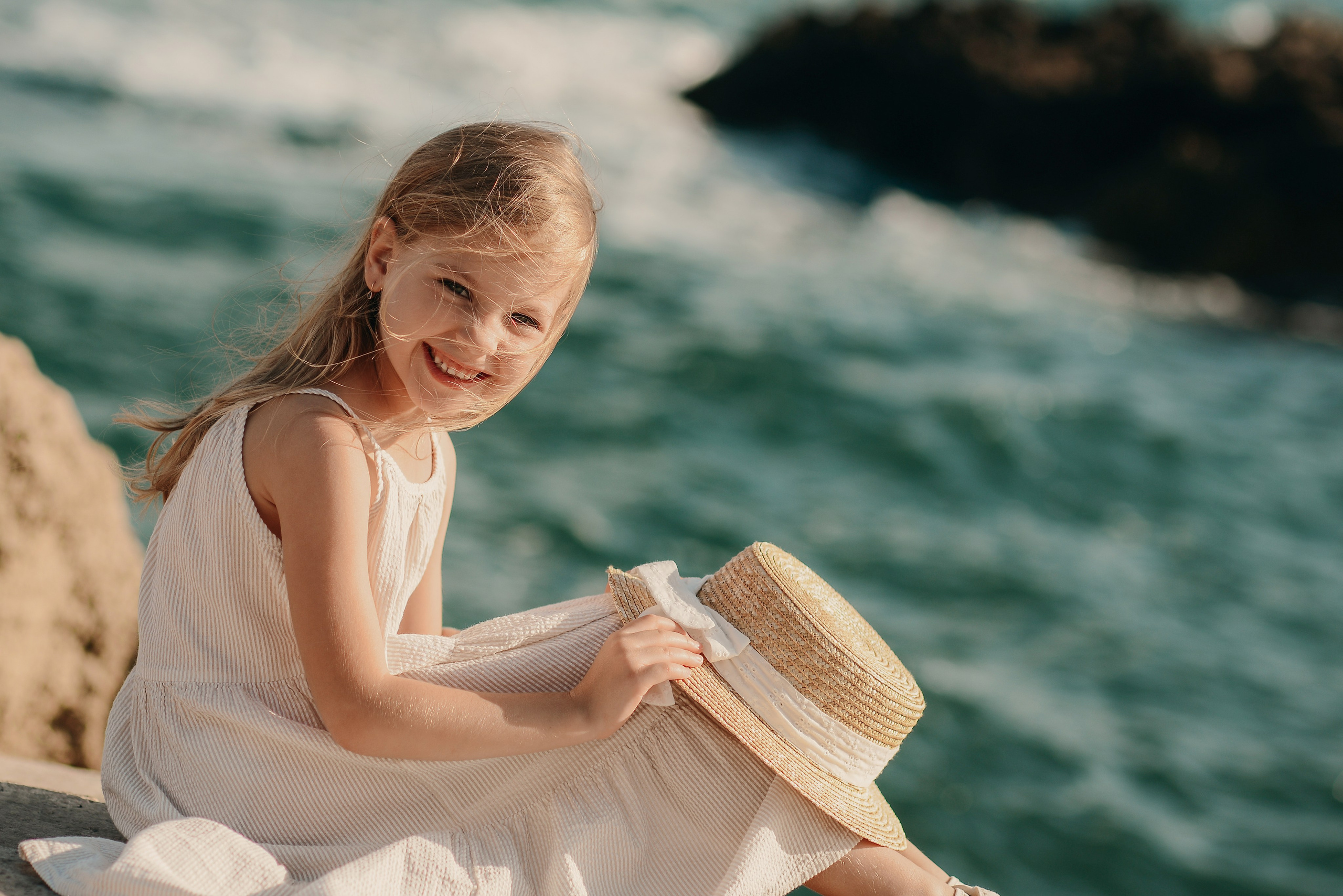 A girl in a light dress sits by the sea in Gallipoli, holding a straw hat and smiling with waves in the background.