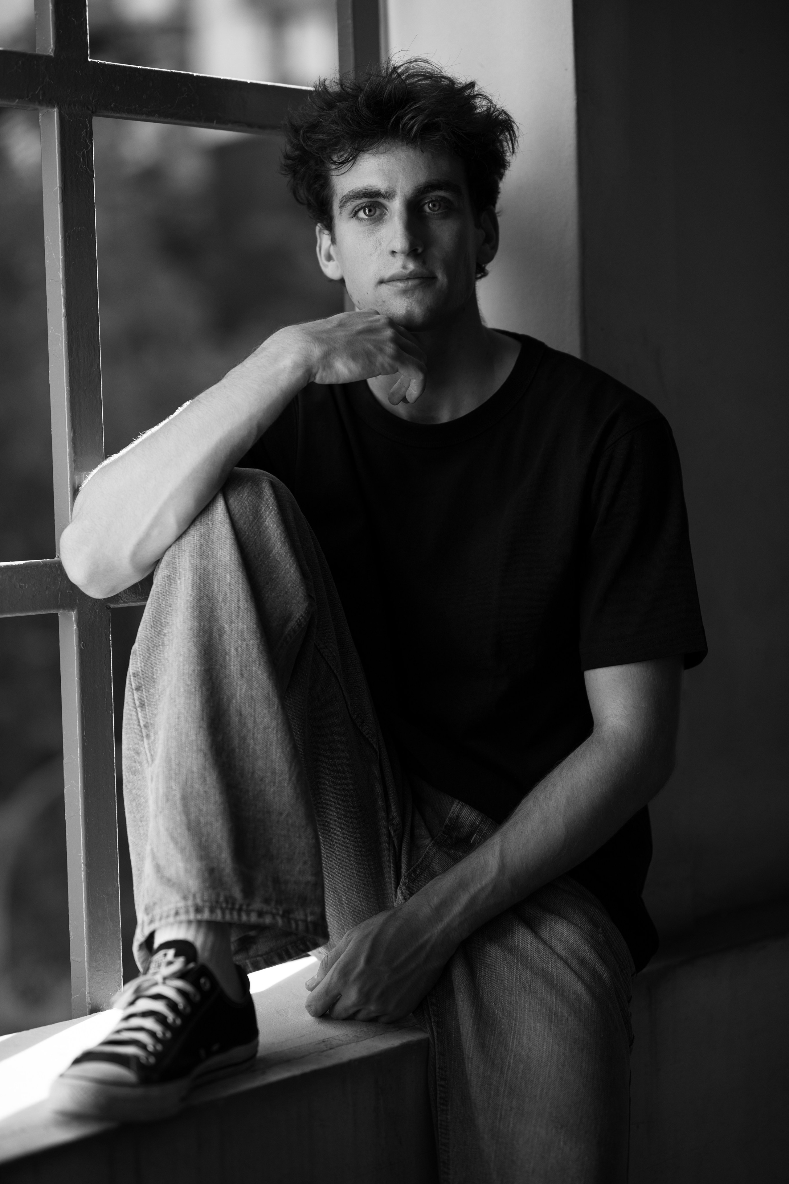 Casual black and white seated portrait of a young man on a windowsill, one knee raised, wearing Converse.