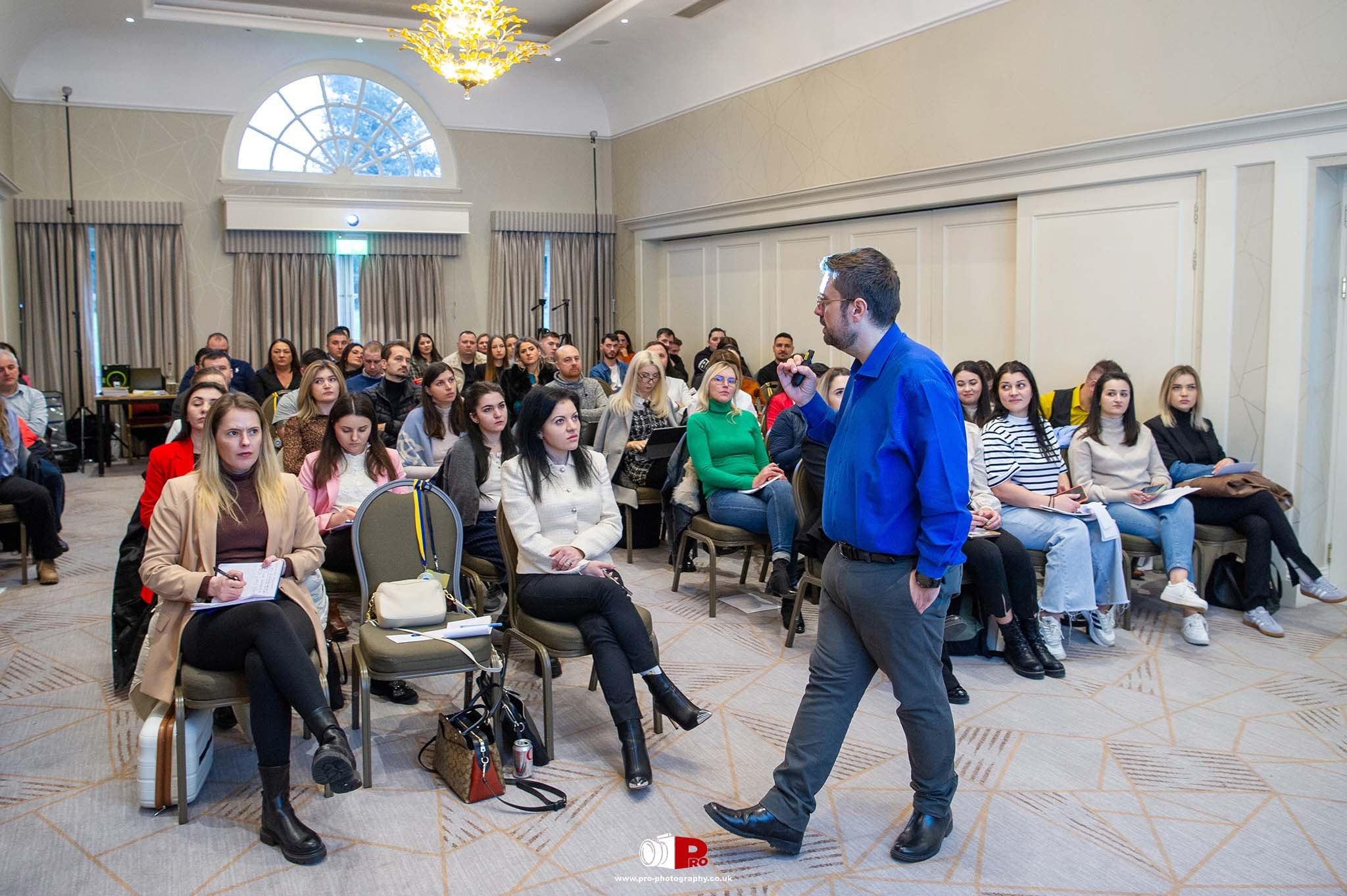 A speaker in a blue shirt addressing a focused audience during a professional seminar in a conference hall.
