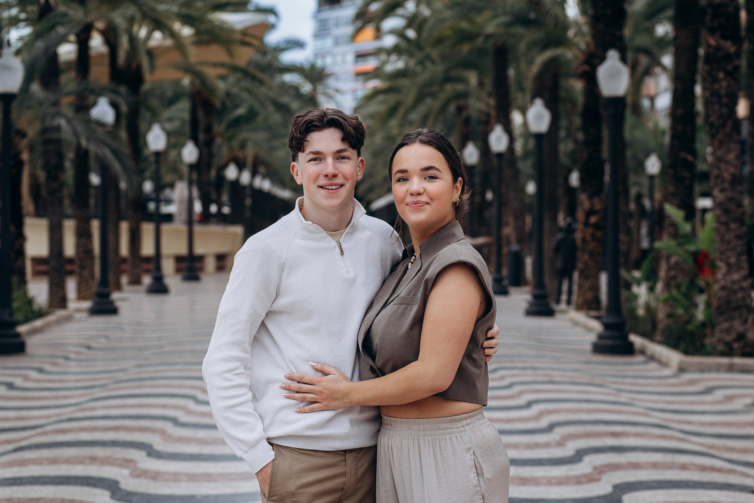 Sesión de fotos familiar en Alicante, España, que muestra un retrato cálido y auténtico de una madre y su hijo adolescente abrazándose en un paseo rodeado de palmeras, capturado con luz natural y arquitectura mediterránea, en una atmósfera urbana relajada — ideal para fotografía familiar, sesiones familiares lifestyle, retratos familiares espontáneos y sesiones familiares emotivas en Alicante y en toda España.