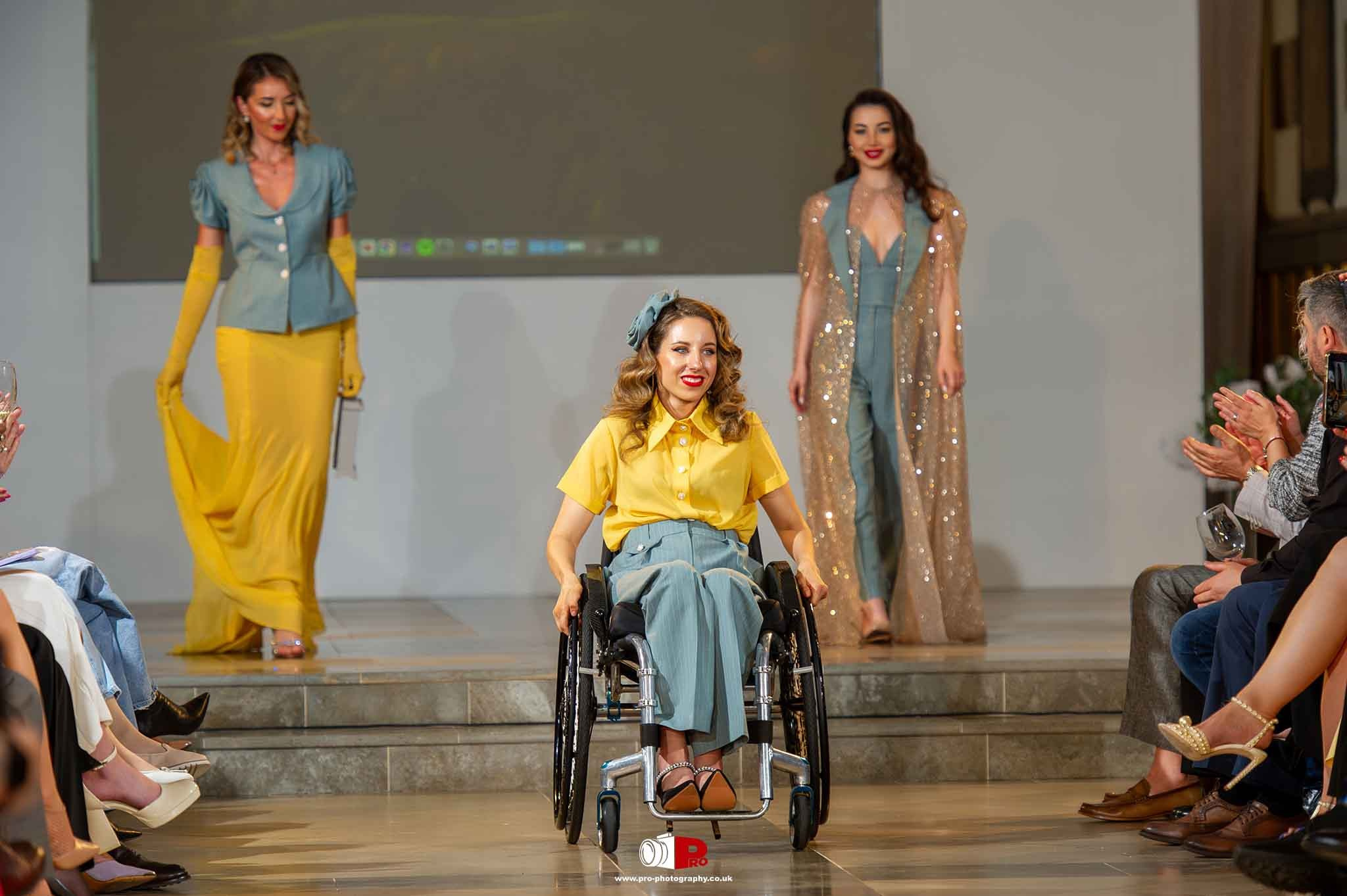 A confident female model in a wheelchair leads the runway, followed by glamorous models in sparkling outfits.