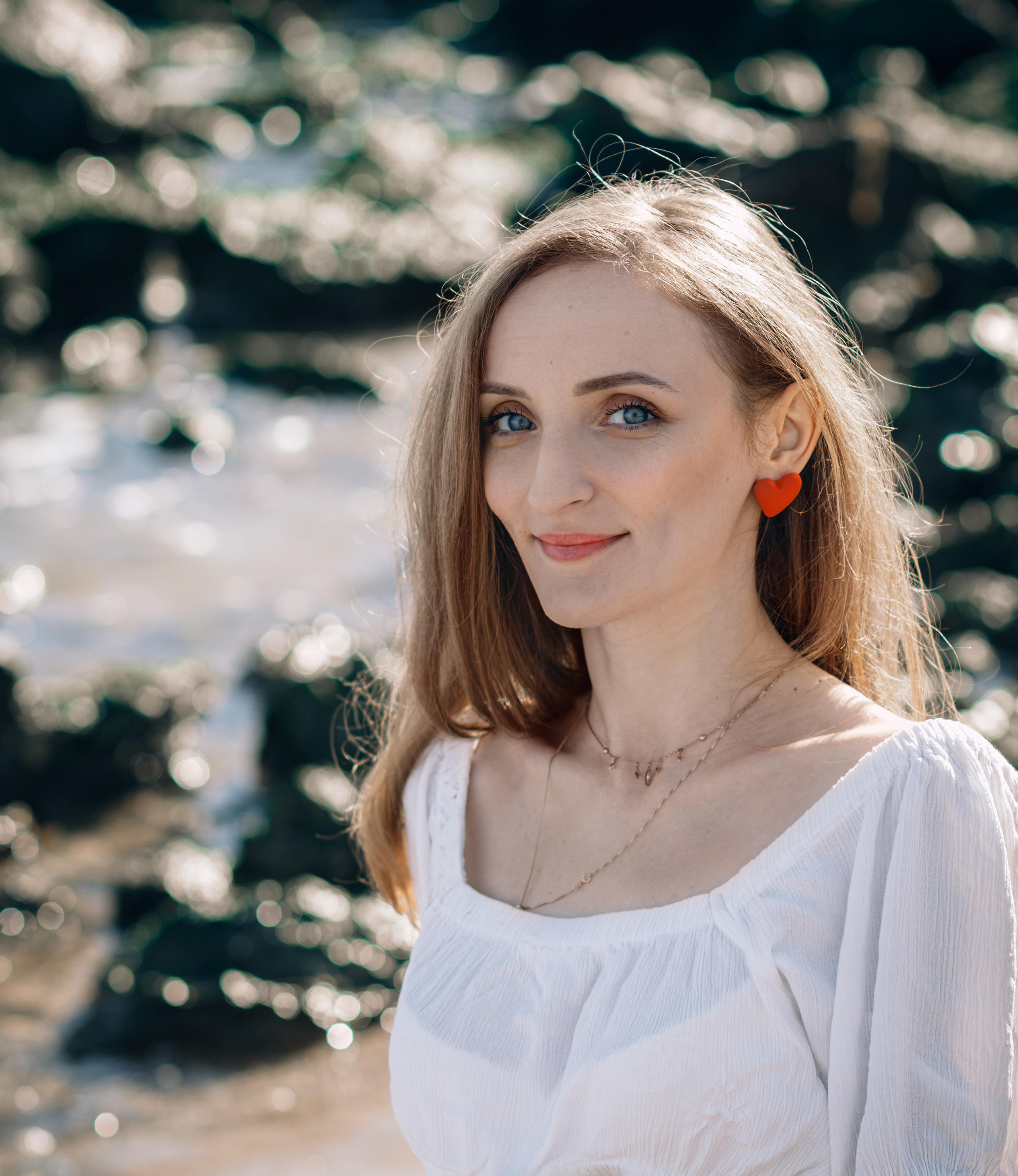 Портрет молодой женщины со светлыми волосами и голубыми глазами, в белой блузке и красных серьгах в форме сердца, с мерцающим морем на заднем плане в Валенсии, Испания