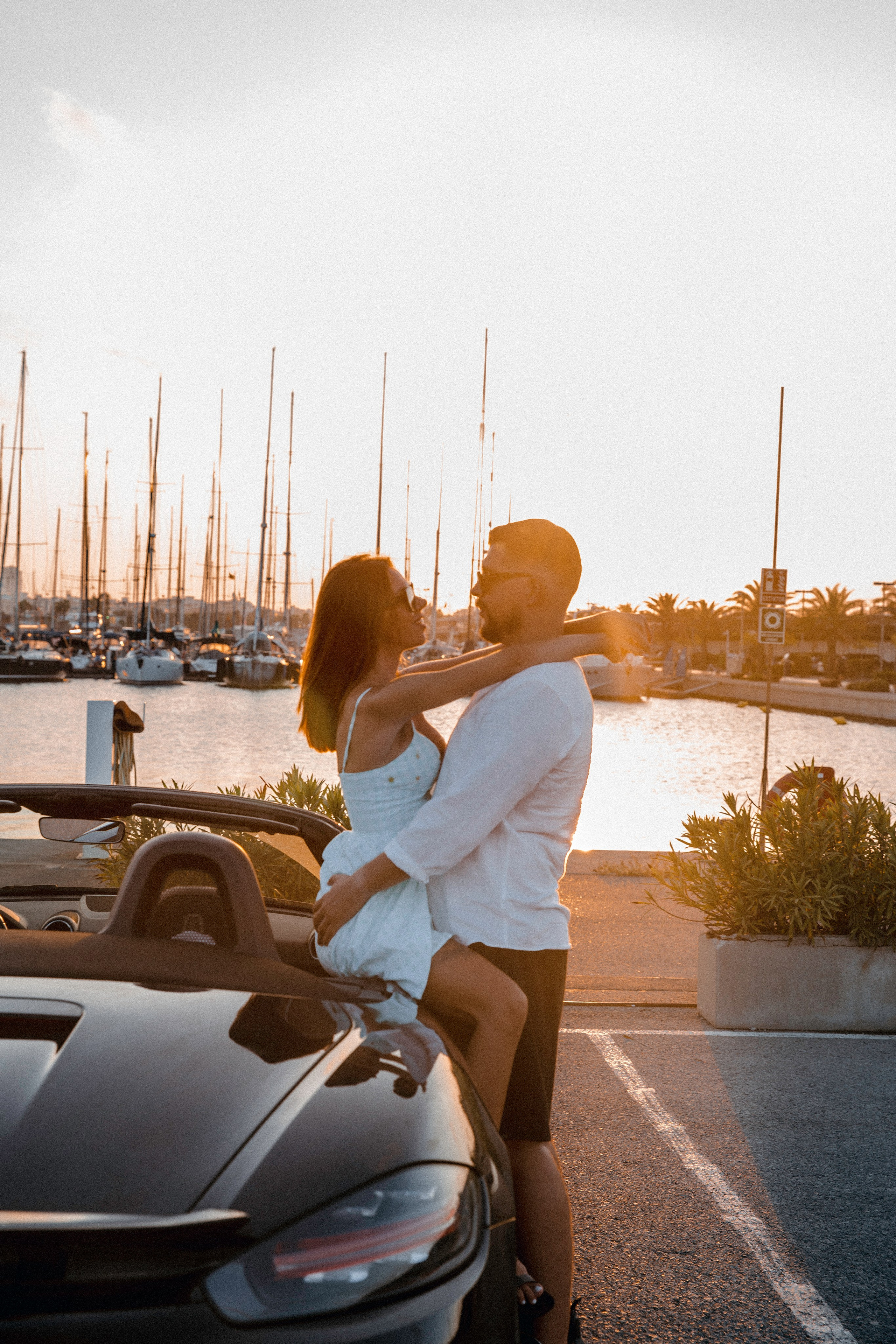Romantic sunset portrait of a couple embracing beside a black convertible at the marina, with yachts and golden light reflecting on the water in the background, capturing a moment of love and connection Valencia, Spain.