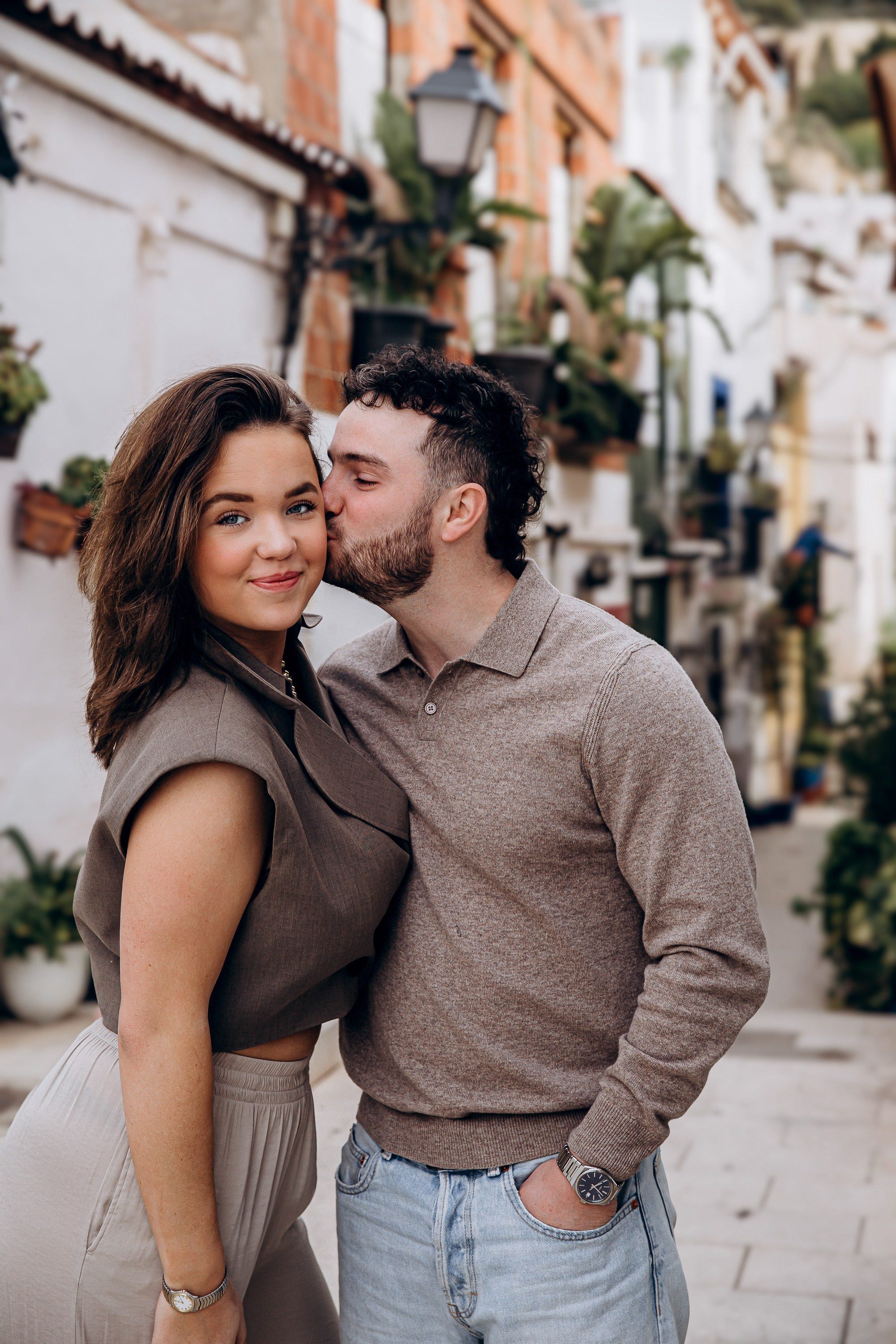 Sesión de fotos love story de pareja en València, España, capturando un momento romántico de una pareja en una encantadora calle histórica, con el hombre besando suavemente la mejilla de su pareja, fotografiado con luz natural suave, arquitectura mediterránea, vegetación y una acogedora atmósfera de casco antiguo — ideal para fotografía de pareja, sesiones love story, reportajes de compromiso, retratos lifestyle románticos y sesiones de pareja en València y en toda España.