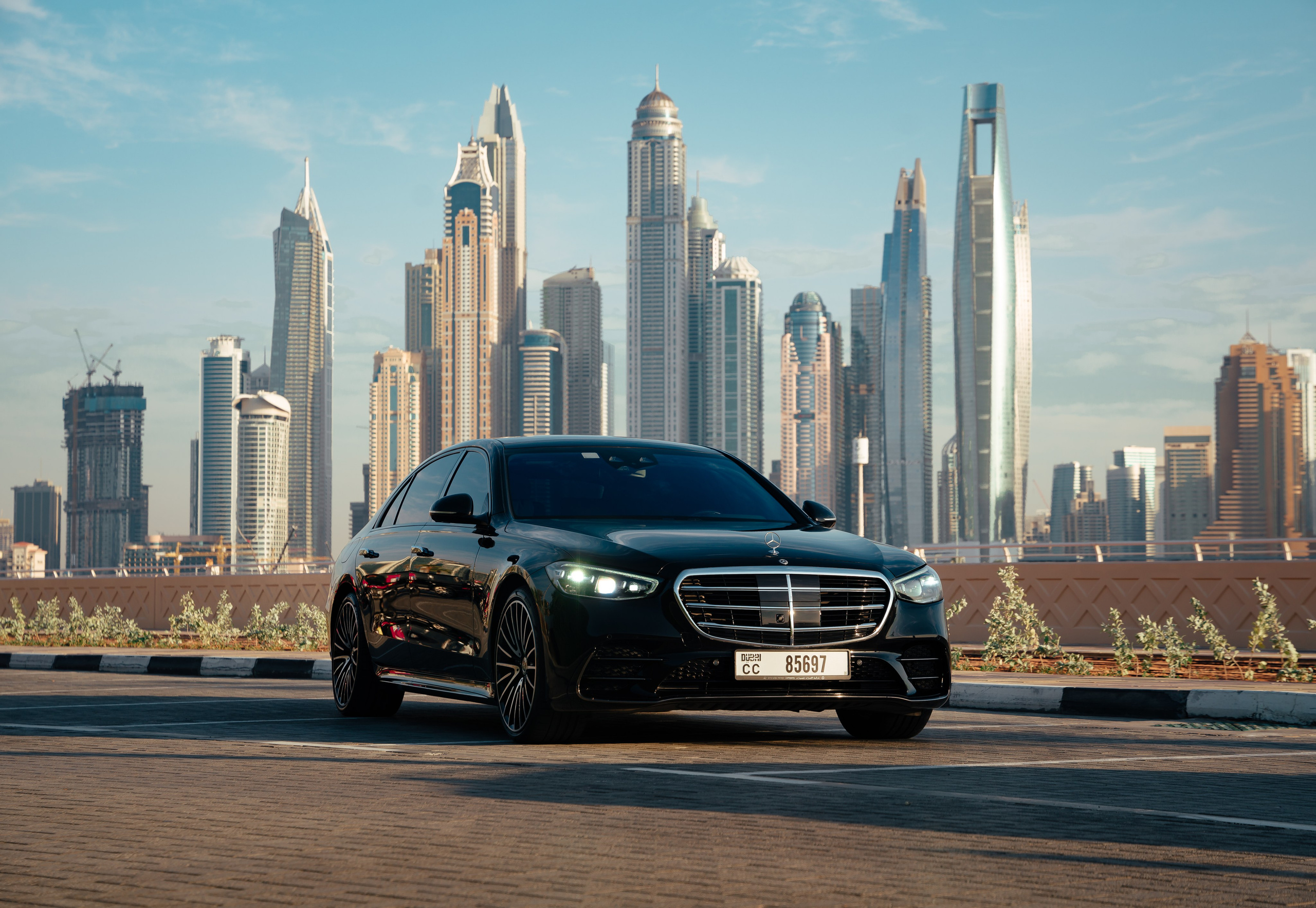 Black Mercedes-Benz with Dubai Marina Skyline: A professional photoshoot captures a sleek black Mercedes-Benz positioned prominently against the backdrop of Dubai’s iconic Marina skyline. The bright, sunny day enhances the vehicle’s polished exterior, highlighting its luxurious design amidst the towering skyscrapers