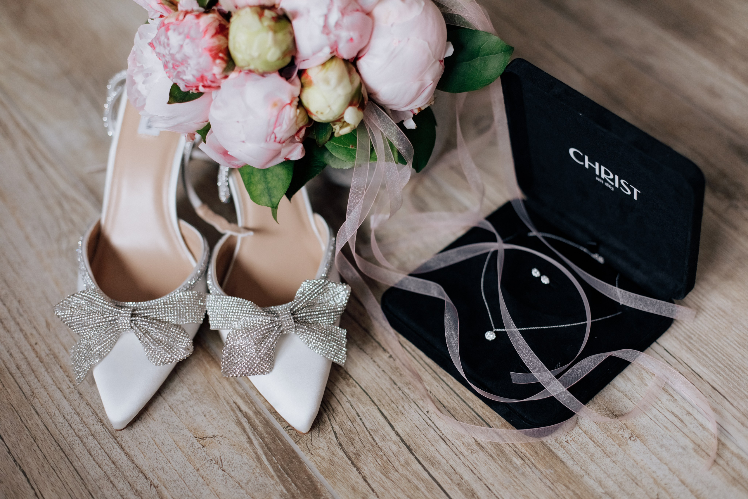 Elegante Brautschuhe mit Schleifen und funkelnden Details neben einer Schmuckschatulle von Christ und Pfingstrosen.