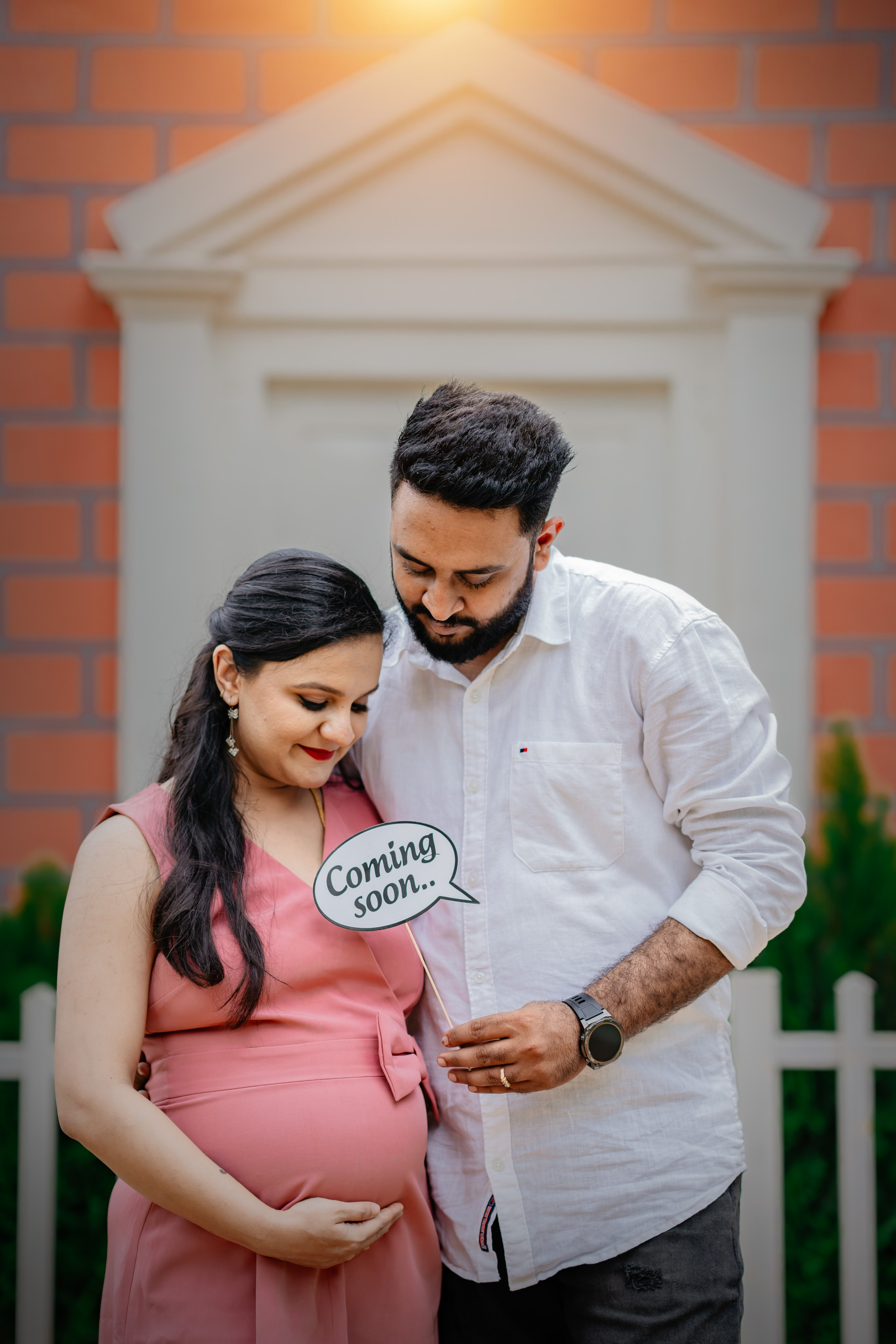 Maternity photoshoot in Bengaluru featuring a man in a white shirt and a woman in a pink dress holding a "Coming soon" sign in front of an orange brick house backdrop.